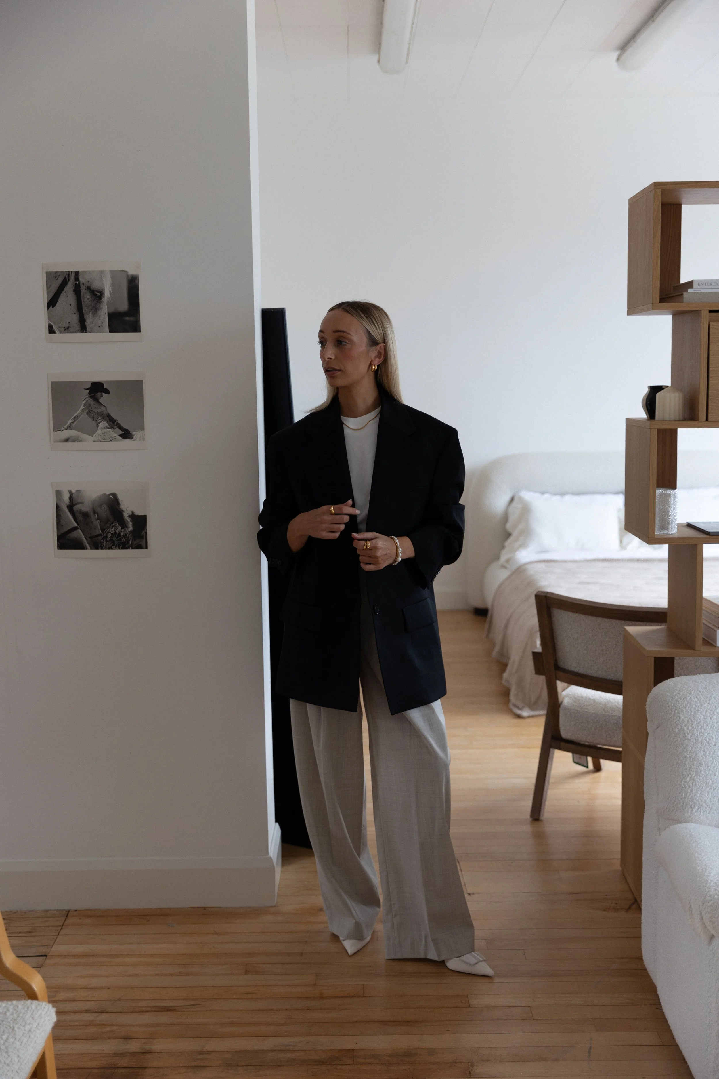 A woman dressed in a black blazer, white top, and light-colored wide-leg trousers standing inside a modern, minimalist room with hardwood floors, white walls, a bed, a chair, and a wooden shelf.