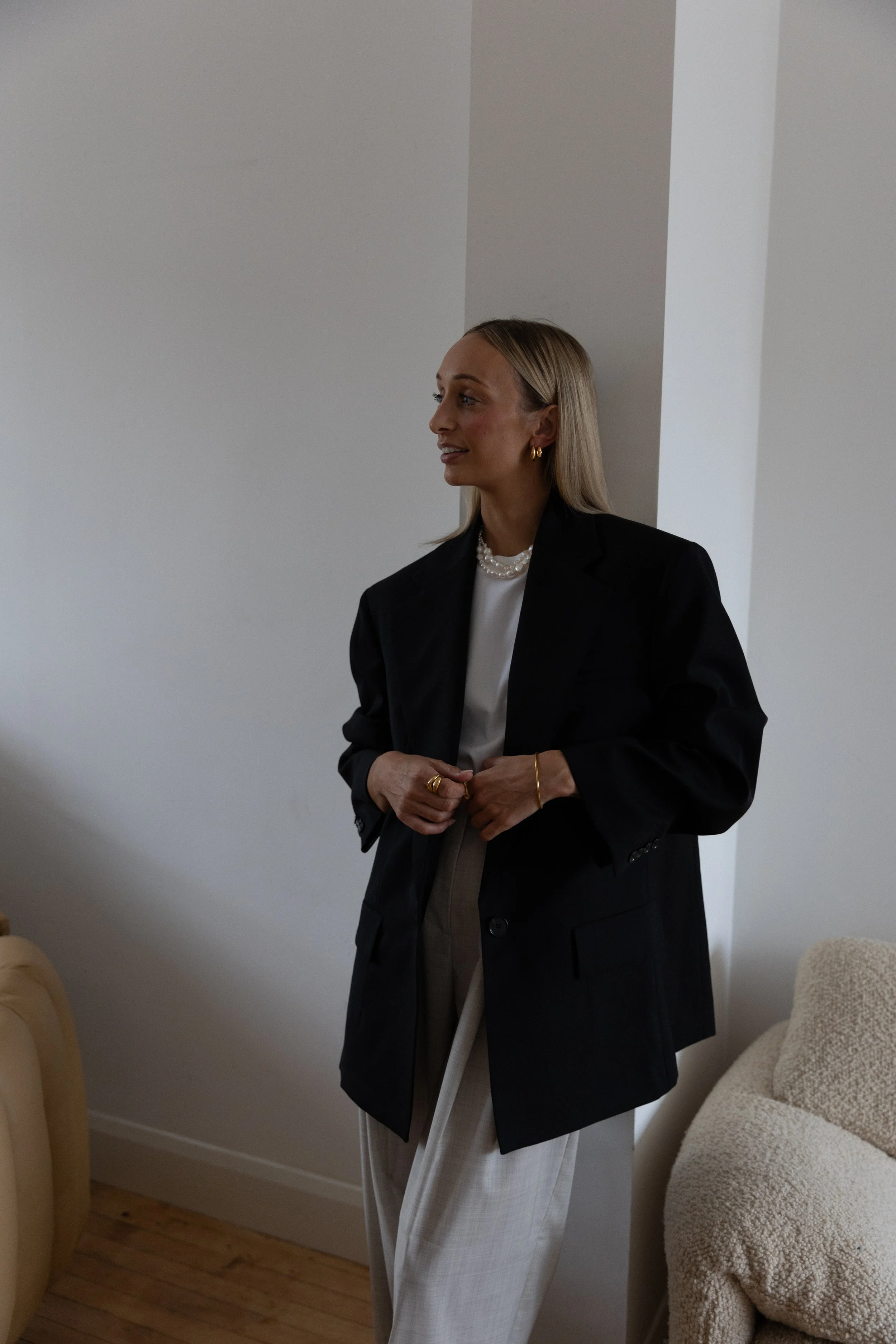 A woman with blonde hair wearing a black blazer, white blouse, and beige trousers standing beside a cream-colored chair. She is accessorized with gold jewelry and has a neutral expression.