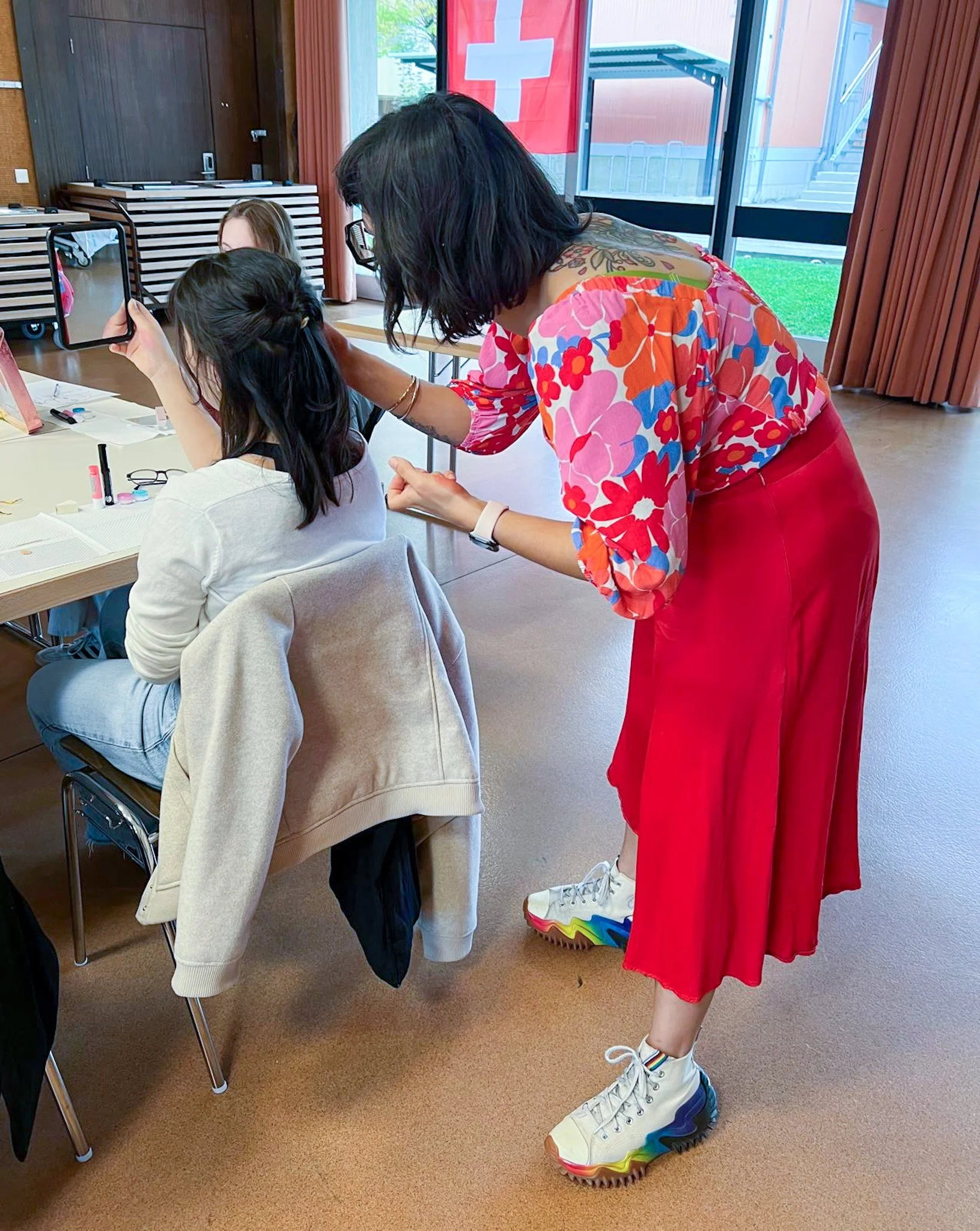 Une femme habillée en rose et rouge aide une autre personne à se faire maquiller ou coiffer lors d'un événement en intérieur avec drapeau suisse en arrière-plan.