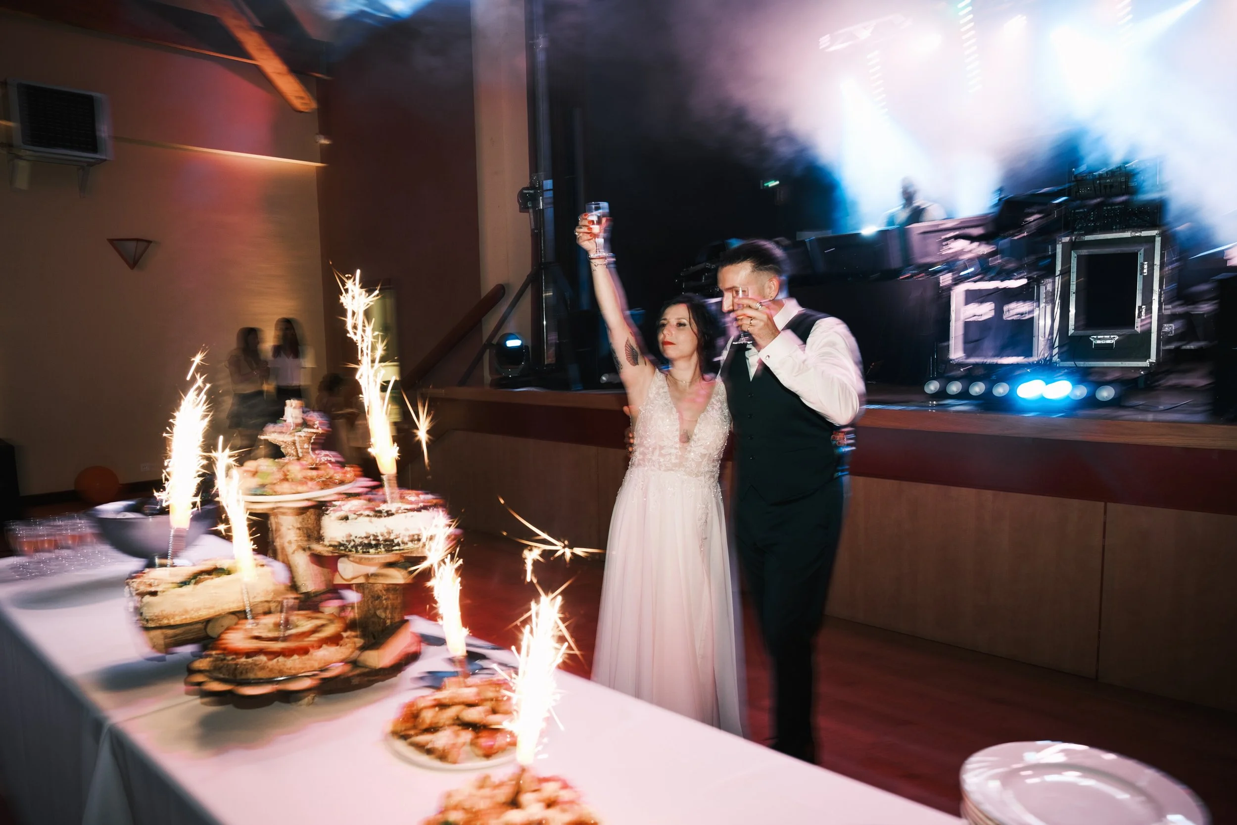 Un couple de mariés célébrant leur mariage lors d'une réception, avec un gâteau d'anniversaire illuminé par des bougies et des feux d'artifice, sur fond d'une scène avec des lumières de fête.
