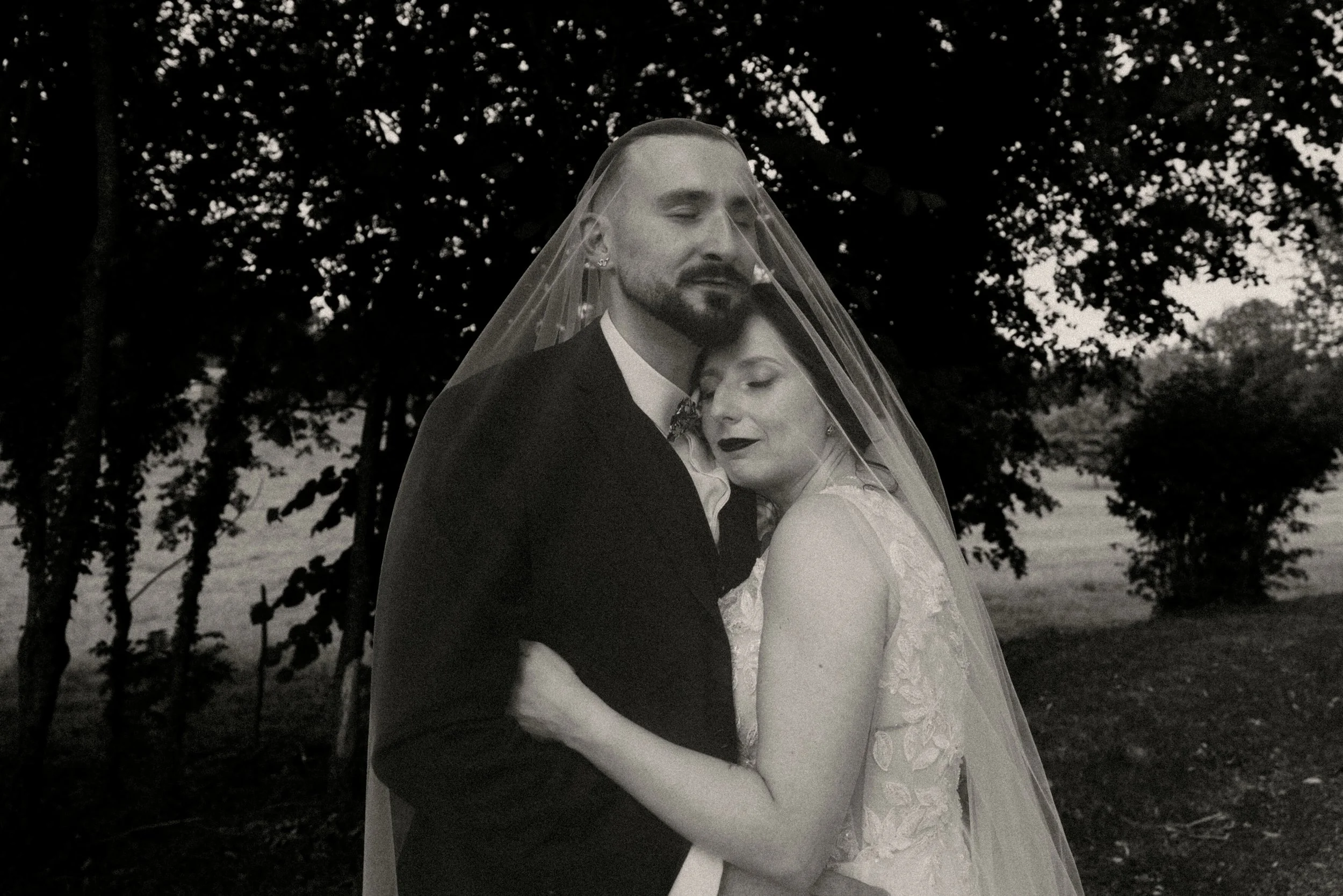 Un couple de mariés dans une clôture en fleurs, avec une femme portant une robe de mariage et un homme en costume, sous un voile, dans un décor extérieur.
