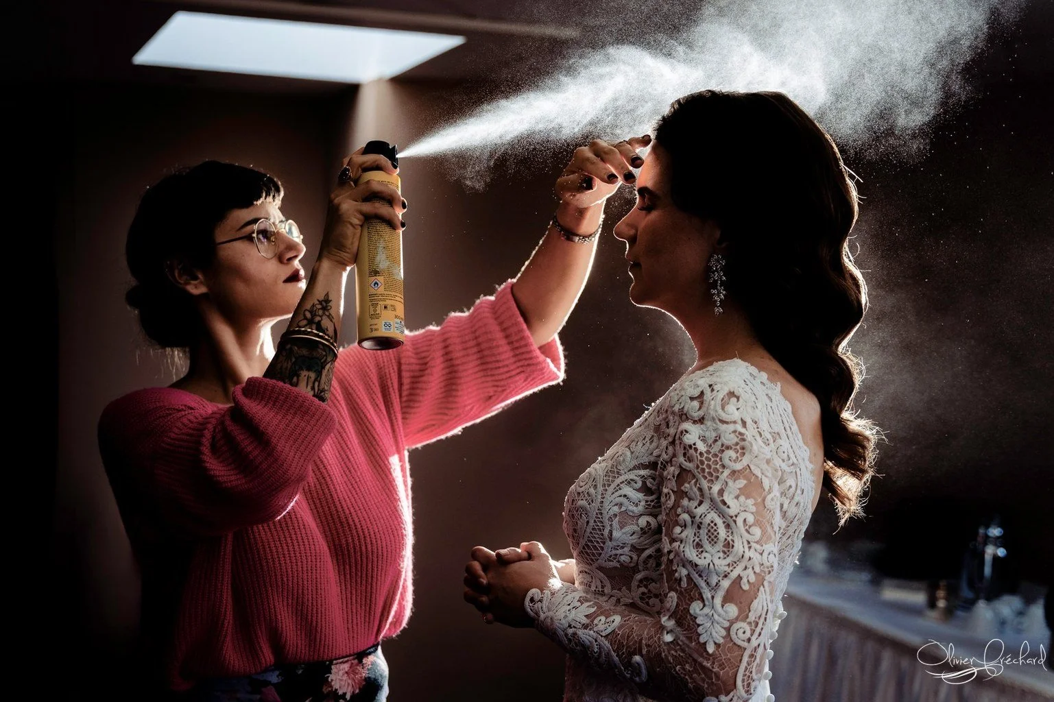 Une femme avec une robe blanche en dentelle se fait asperger de poudre par une autre femme portant un pull rose, lors d'une préparation pour un mariage.