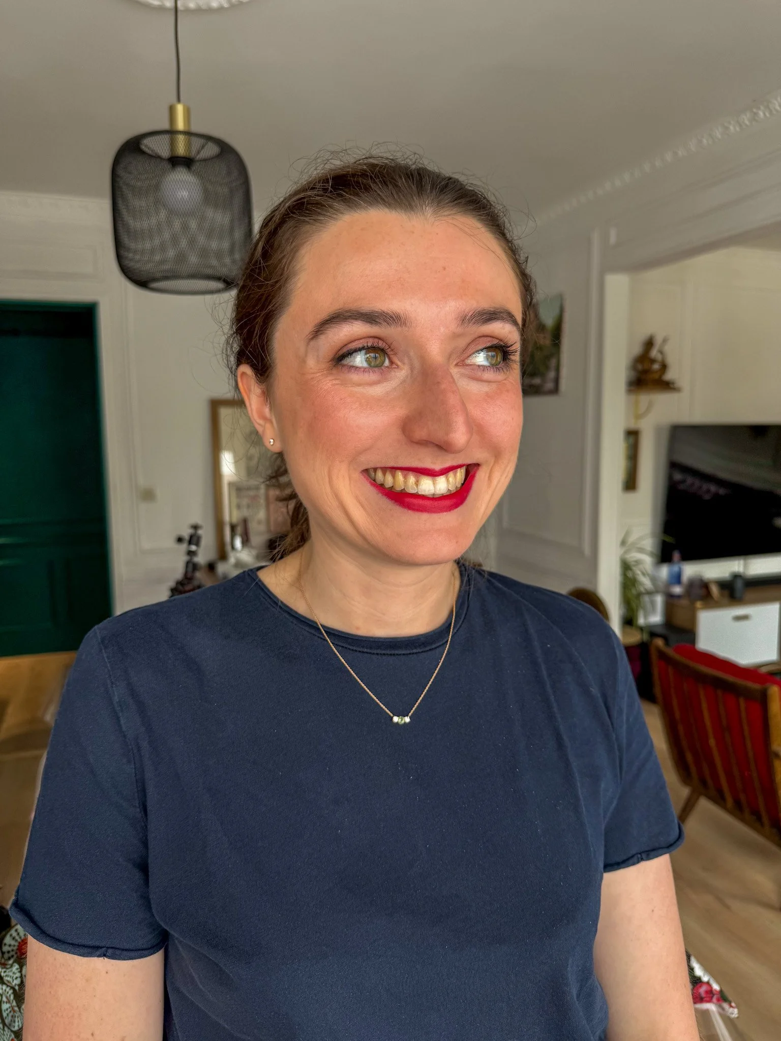 Une femme souriante avec des cheveux attachés, portant un t-shirt bleu foncé et un collier, se trouve dans un intérieur résidentiel lumineux avec un téléviseur, un bureau, et des décorations murales.