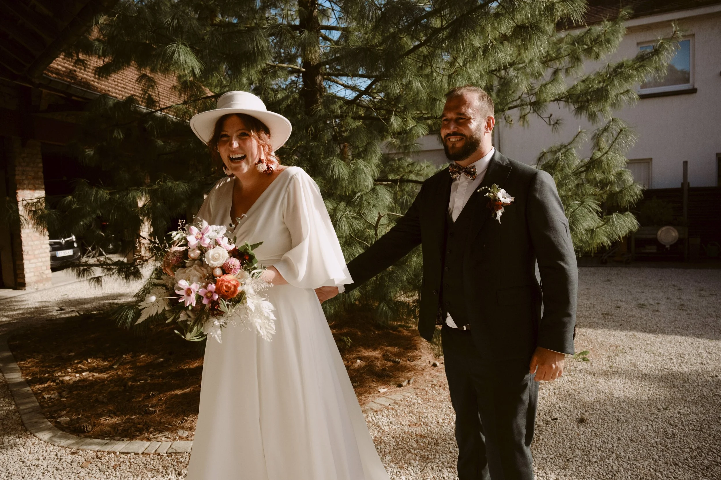 Un couple marié souriant, la femme en robe blanche et un chapeau, tenant un bouquet de fleurs, l'homme en smoking avec un nœud papillon à côté d'un arbre vert.