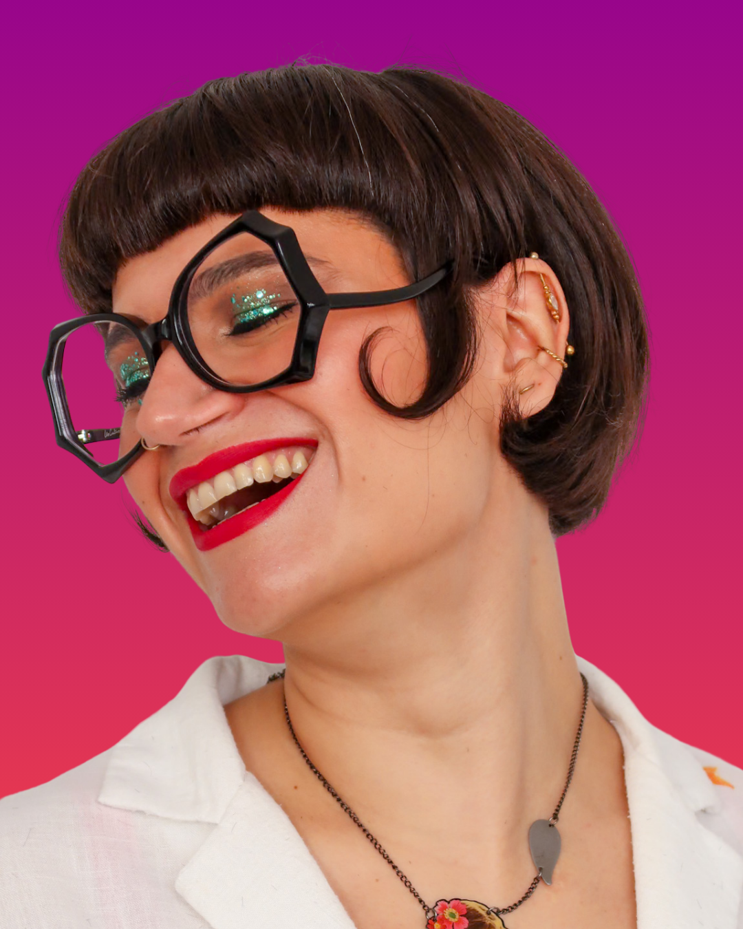 une femme souriante avec des lunettes noires de forme géométrique, des lèvres rouges, des boucles d'oreilles et un collier, contre un fond violet et rose dégradé.