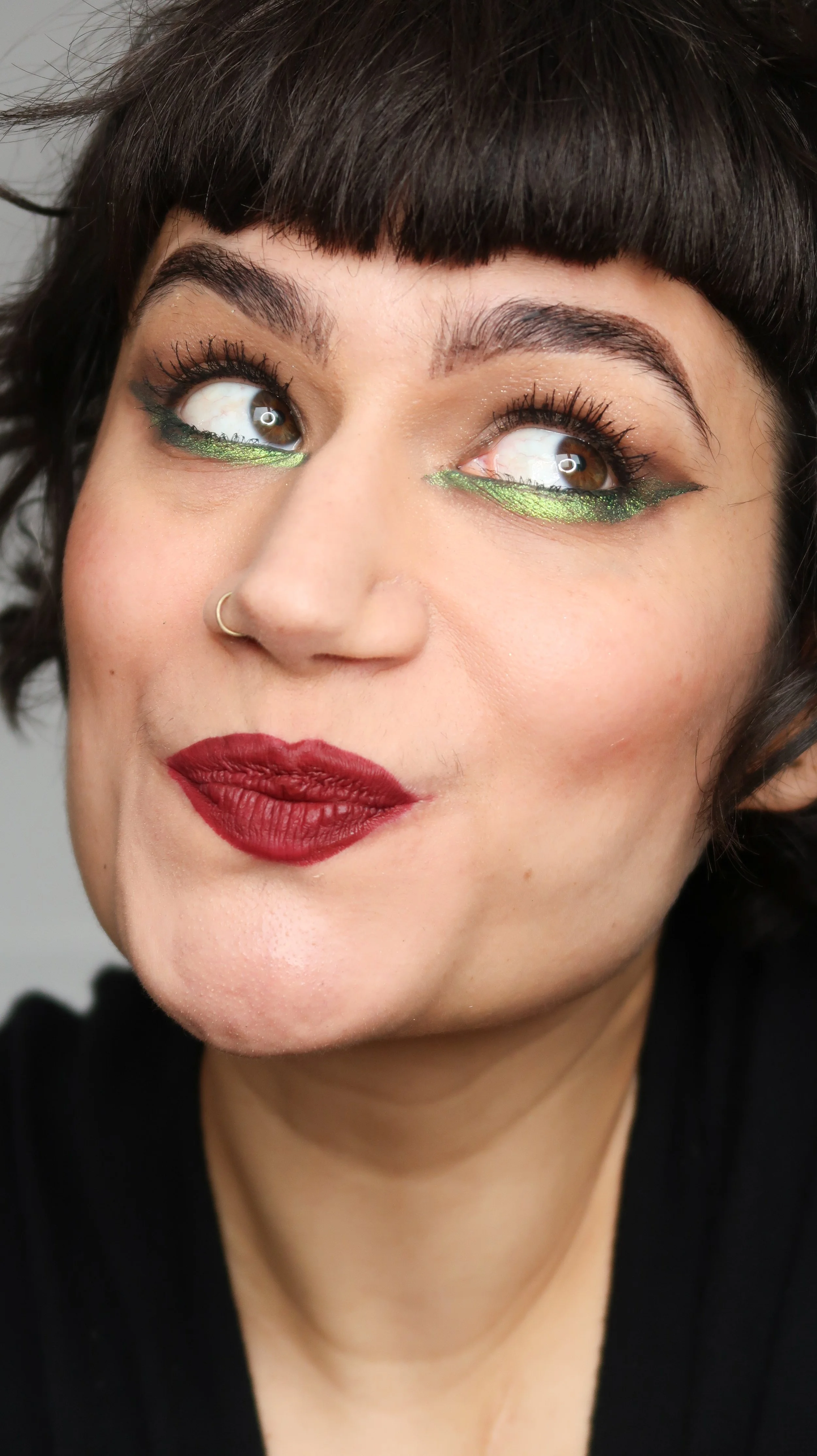 Une femme avec des yeux maquillés avec un eye-liner vert brillant, des lèvres rouge foncé, et des cheveux courts et bouclés.