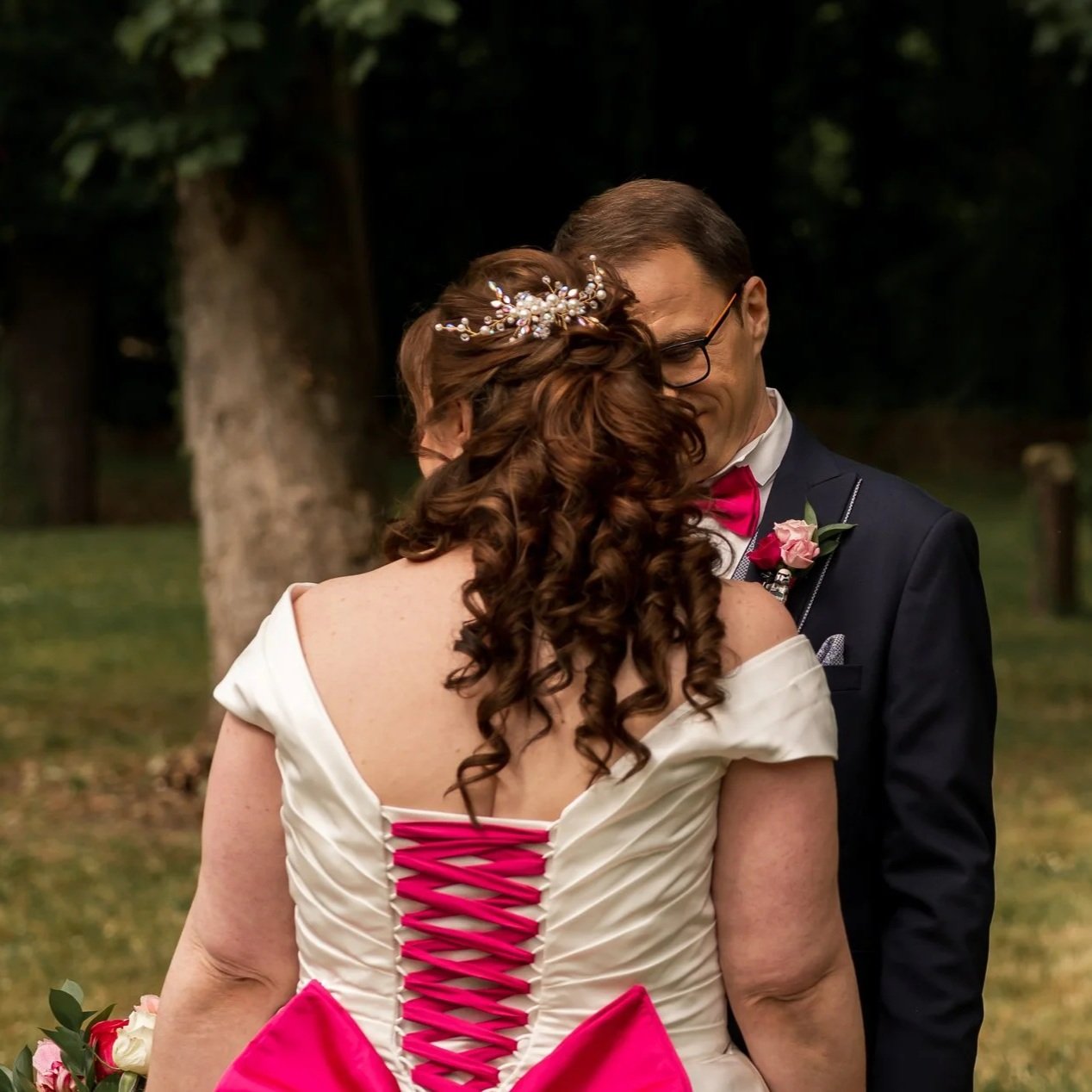 Un couple de mariés lors d'une cérémonie en plein air, la femme a des cheveux bouclés avec une décoration florale dans les cheveux, portant une robe blanche avec un dos lacé rose, l'homme porte un costume sombre avec une chemise blanche et un noeud papillon rose.