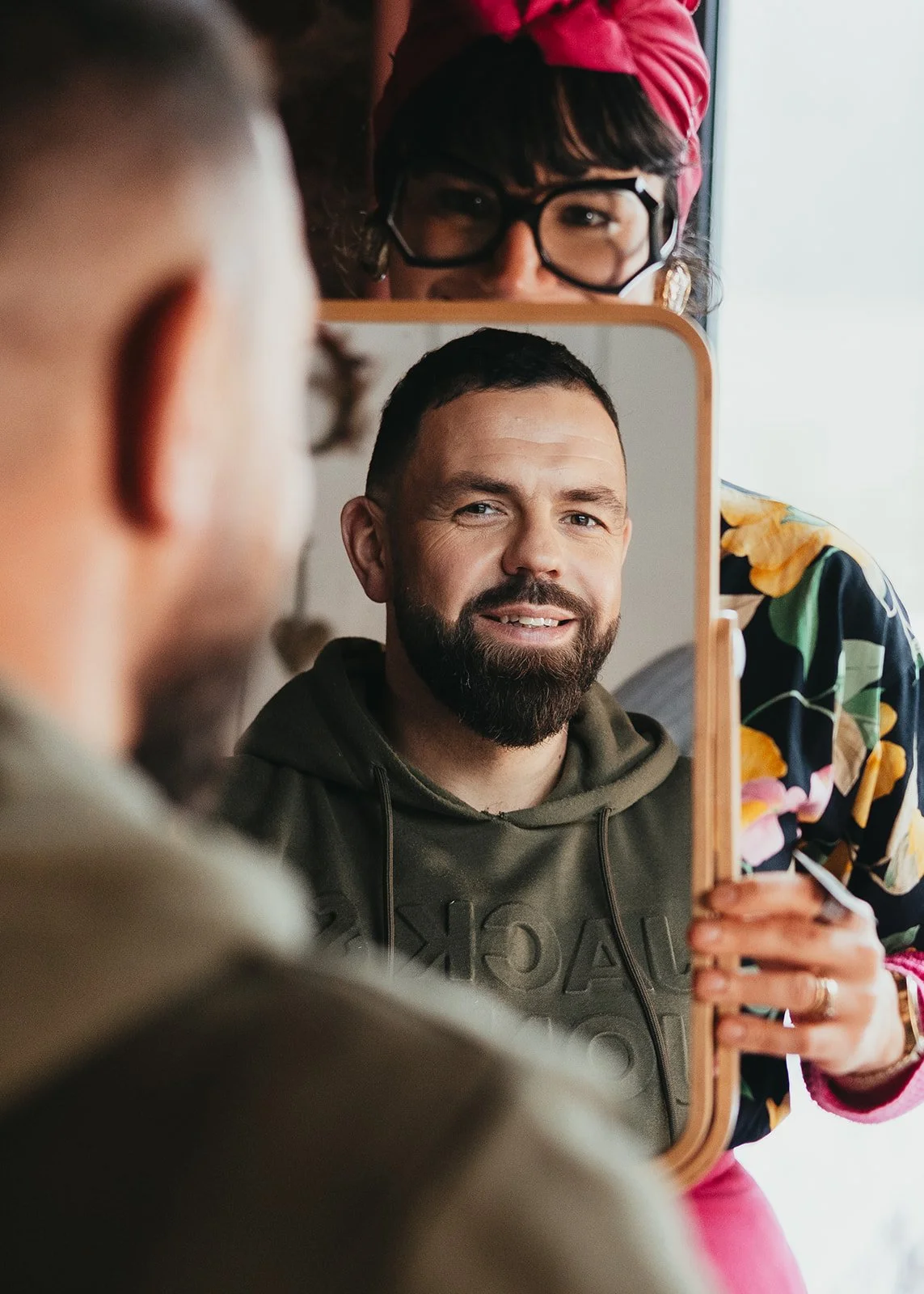 Une femme avec un foulard rose et des lunettes regarde dans un miroir, en face d'un homme aux cheveux courts et à la barbe, qui se regarde dans le miroir.