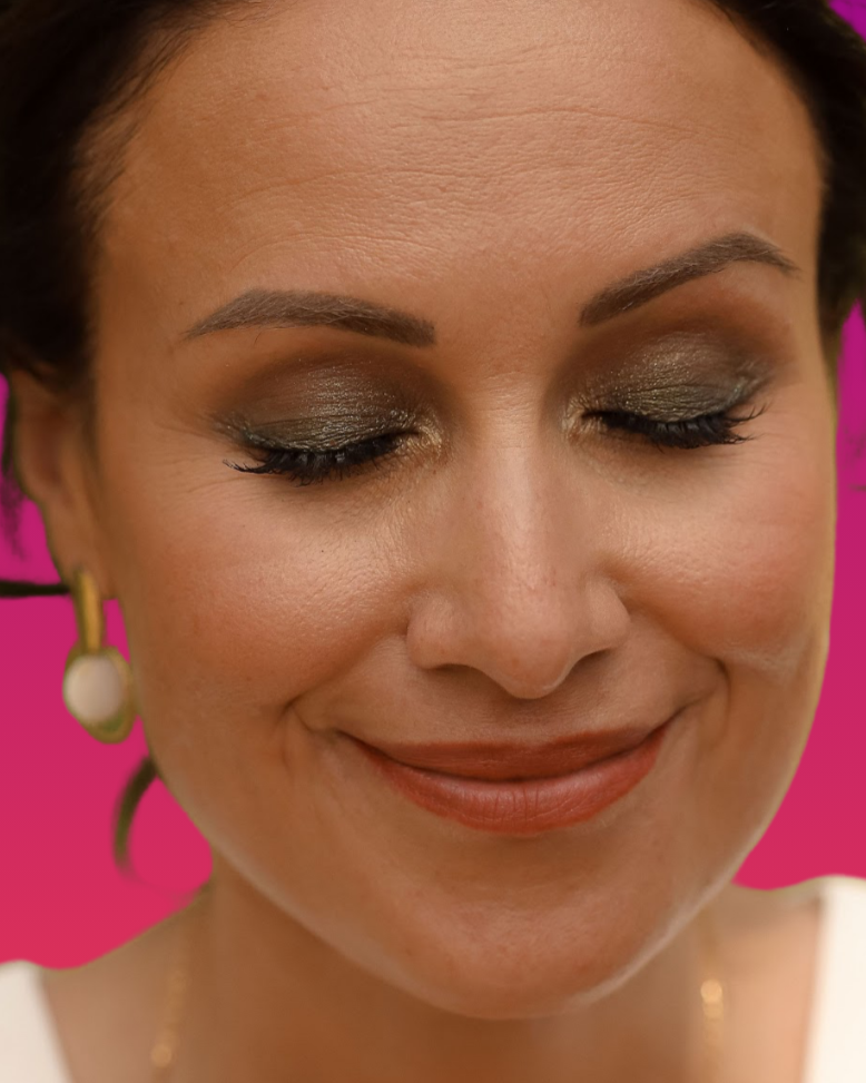 Close-up d'un visage de femme avec maquillage sophistiqué, souriant doucement, portant des boucles d'oreilles et un maquillage en ton doré et vert, sur un fond rose.