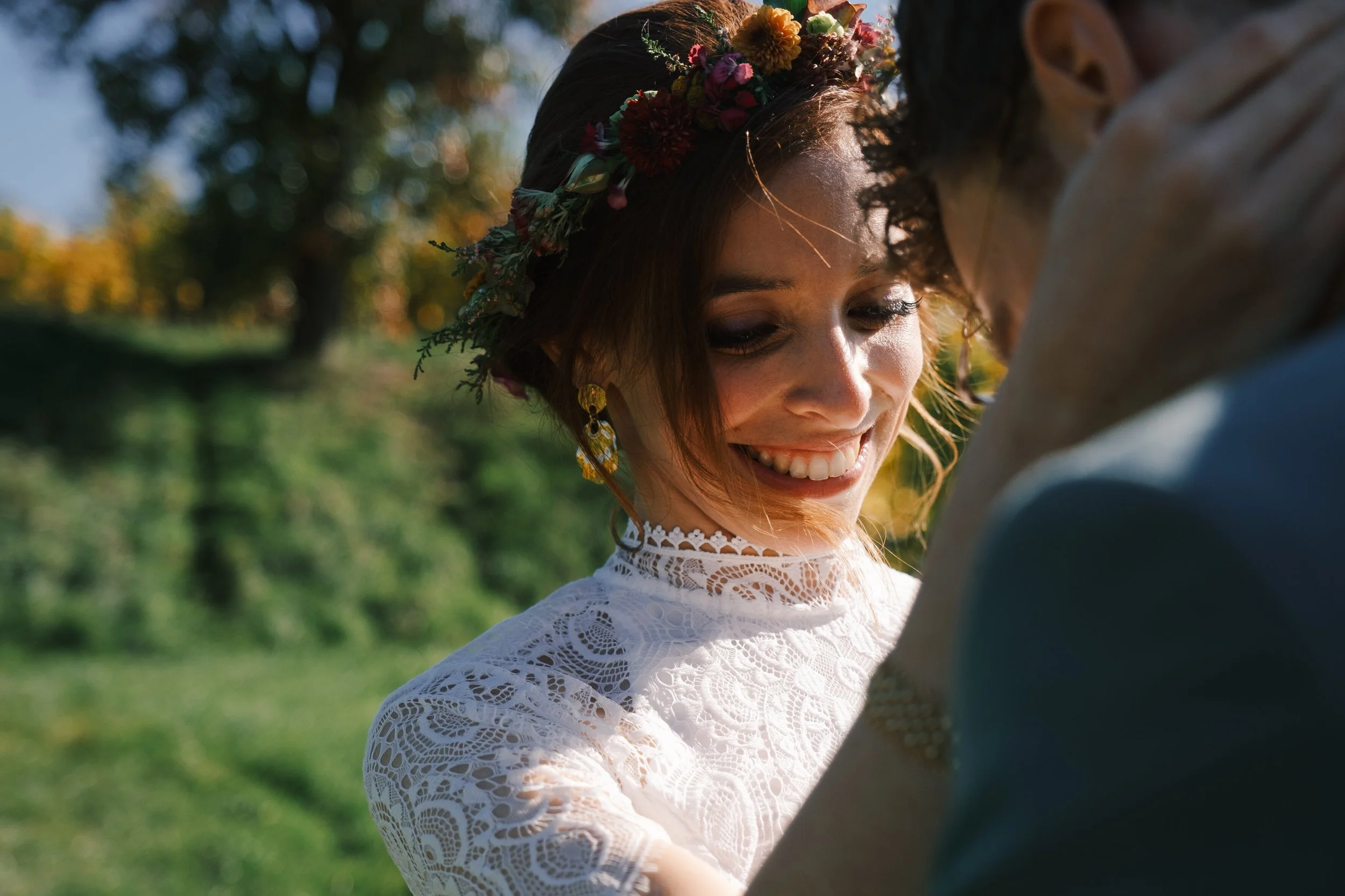 Un couple souriant lors d'un mariage en plein air, la femme porte une robe en dentelle et un couronne de fleurs, la scène est en plein jour avec un fond naturel verdoyant.