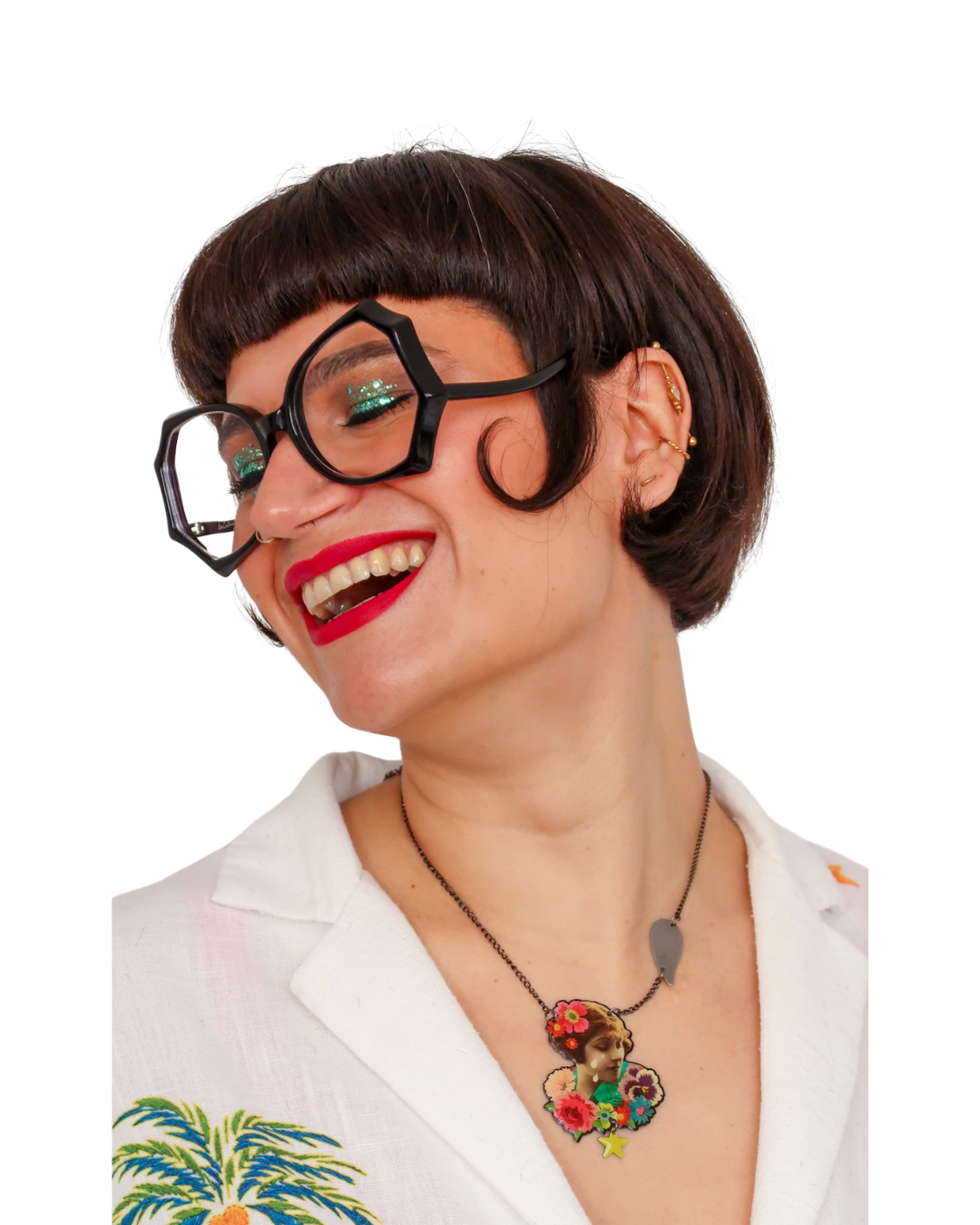 Une femme souriante, portant des lunettes épaisses, un collier avec une femme en pendentif et une chemise blanche avec des broderies colorées.