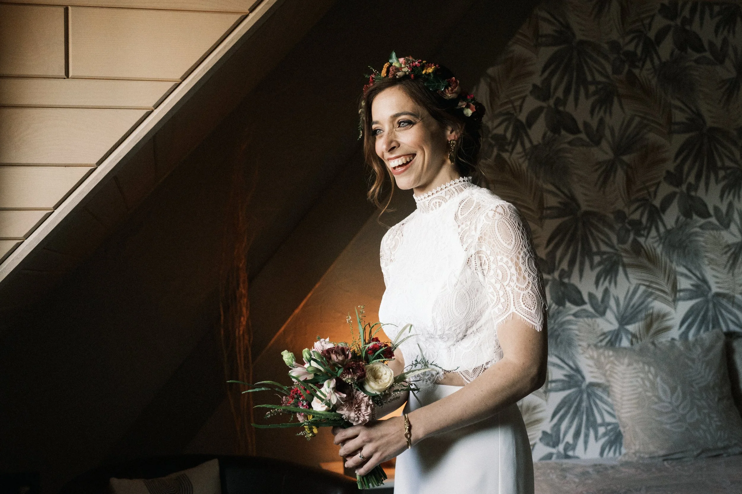 une femme souriante en robe blanche, portant une couronne de fleurs, tenant un bouquet de fleurs, dans une pièce décorée avec un mur à motifs végétaux