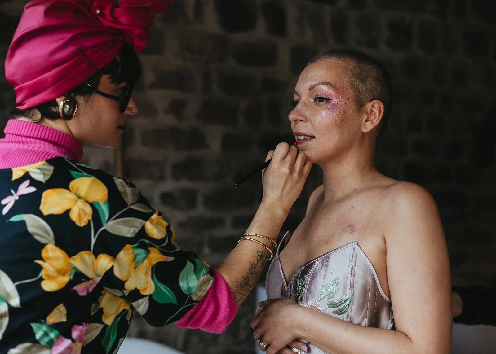 Une femme peint le maquillage d'une autre femme. La femme qui se fait maquiller a des cheveux rasés et porte une robe en satin avec des motifs floraux. La maquilleuse porte un turban rose et un haut à motifs floraux colorés.