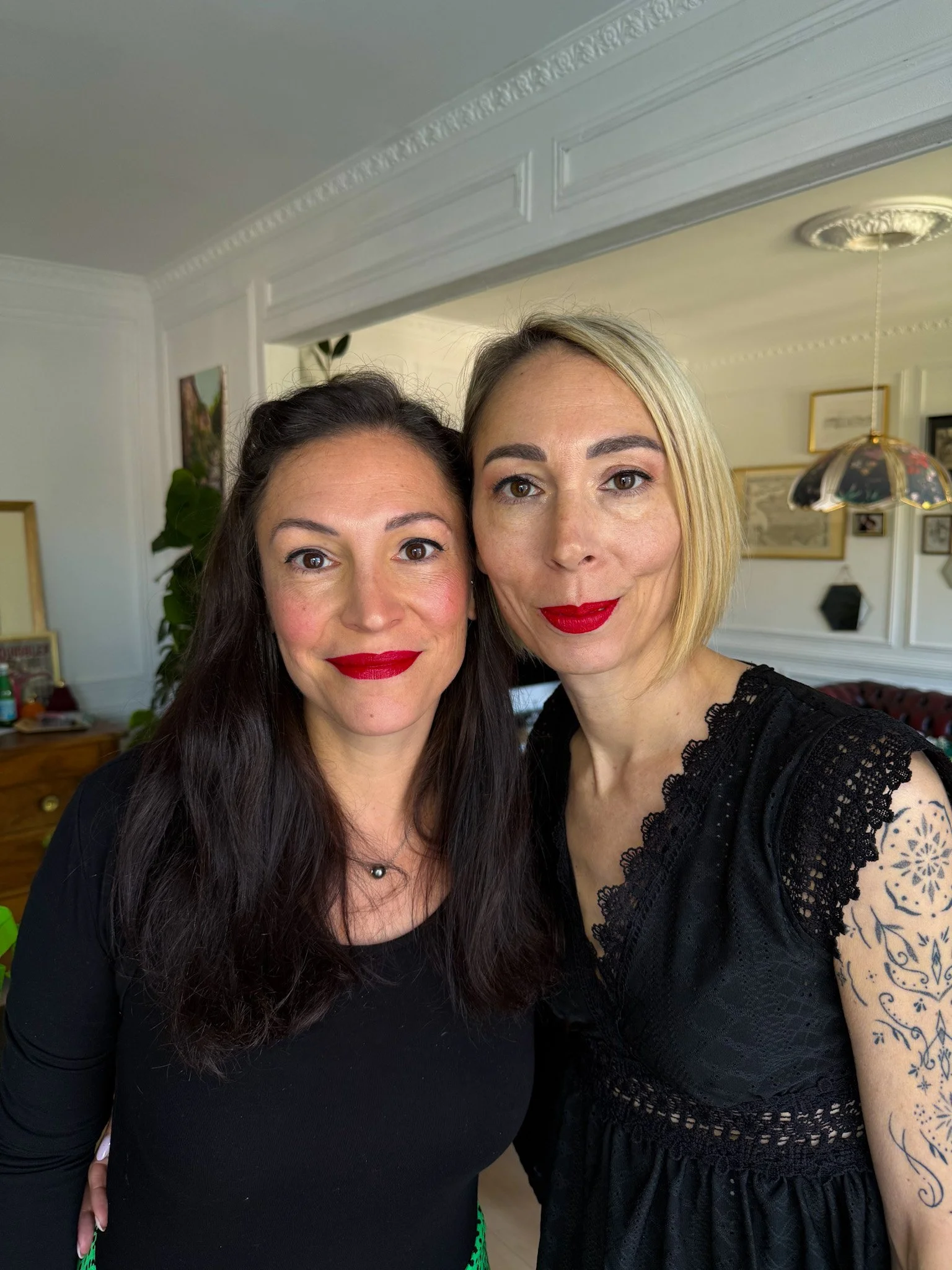 Deux femmes posant pour une photo, toutes deux portant du rouge à lèvres rouge, dans une pièce décorée de cadres et d’un lampadaire suspendu.