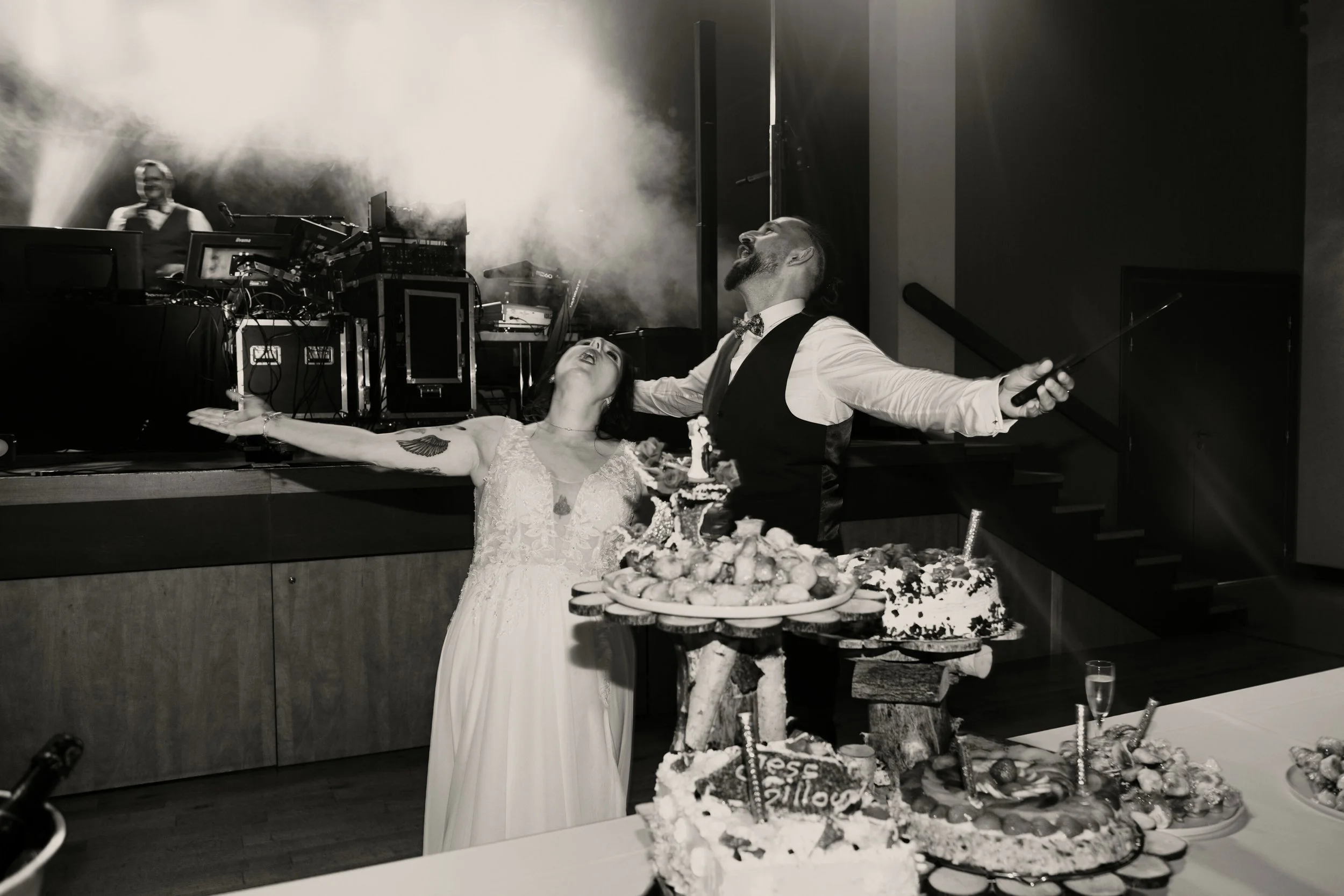Un couple lors d'un mariage, la femme en robe blanche et l'homme en costume, autour d'un gâteau décoré, avec un DJ en arrière-plan dans une ambiance festive.
