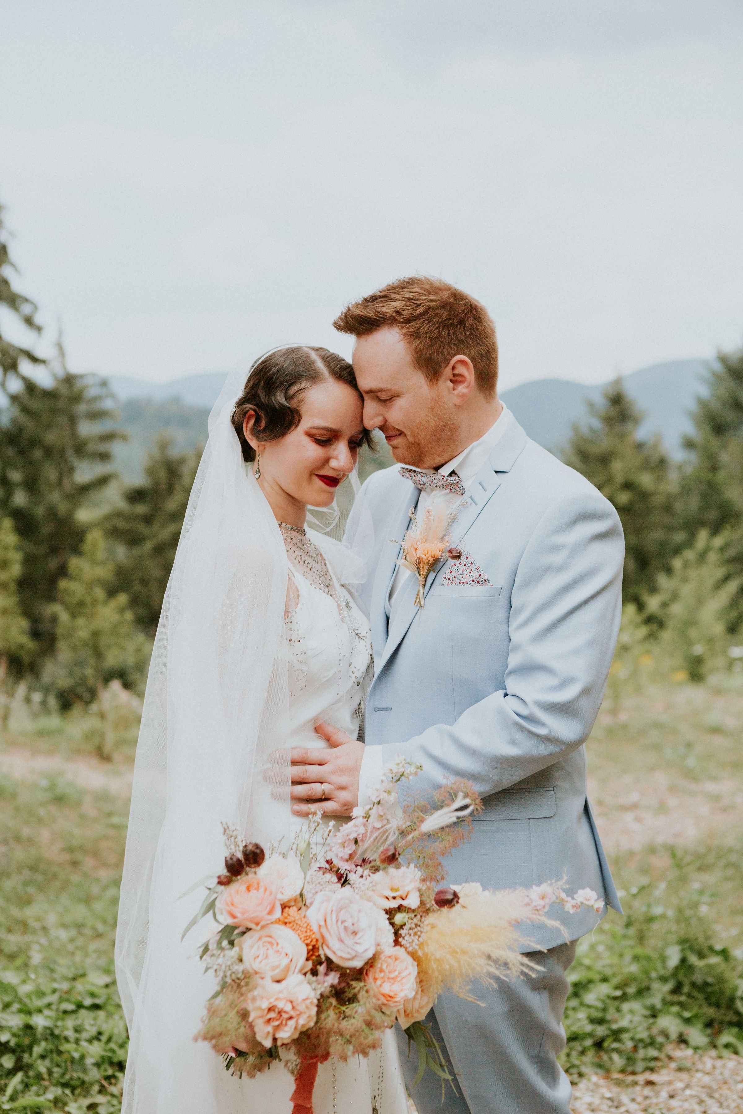 Un couple de mariés partage un moment intime en extérieur, la femme porte une robe blanche avec un voile et tient un bouquet de fleurs, l'homme en costume bleu clair. Ils se touchent le front et ferment les yeux, entourés d'arbres et de montagnes en arrière-plan.
