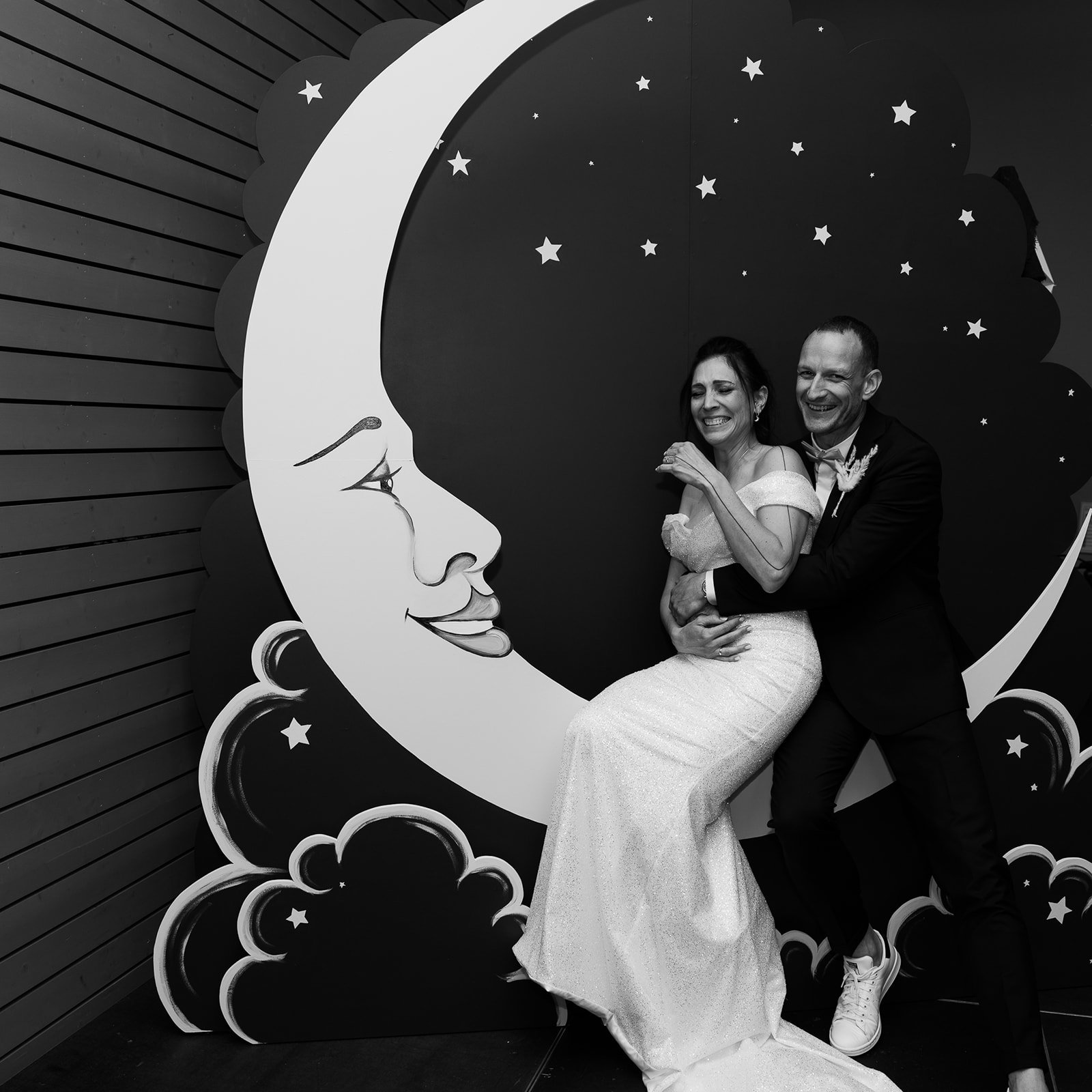 Un couple en robe de mariée et costume posant devant une décoration de lune et étoiles dans un style illustré noir et blanc.