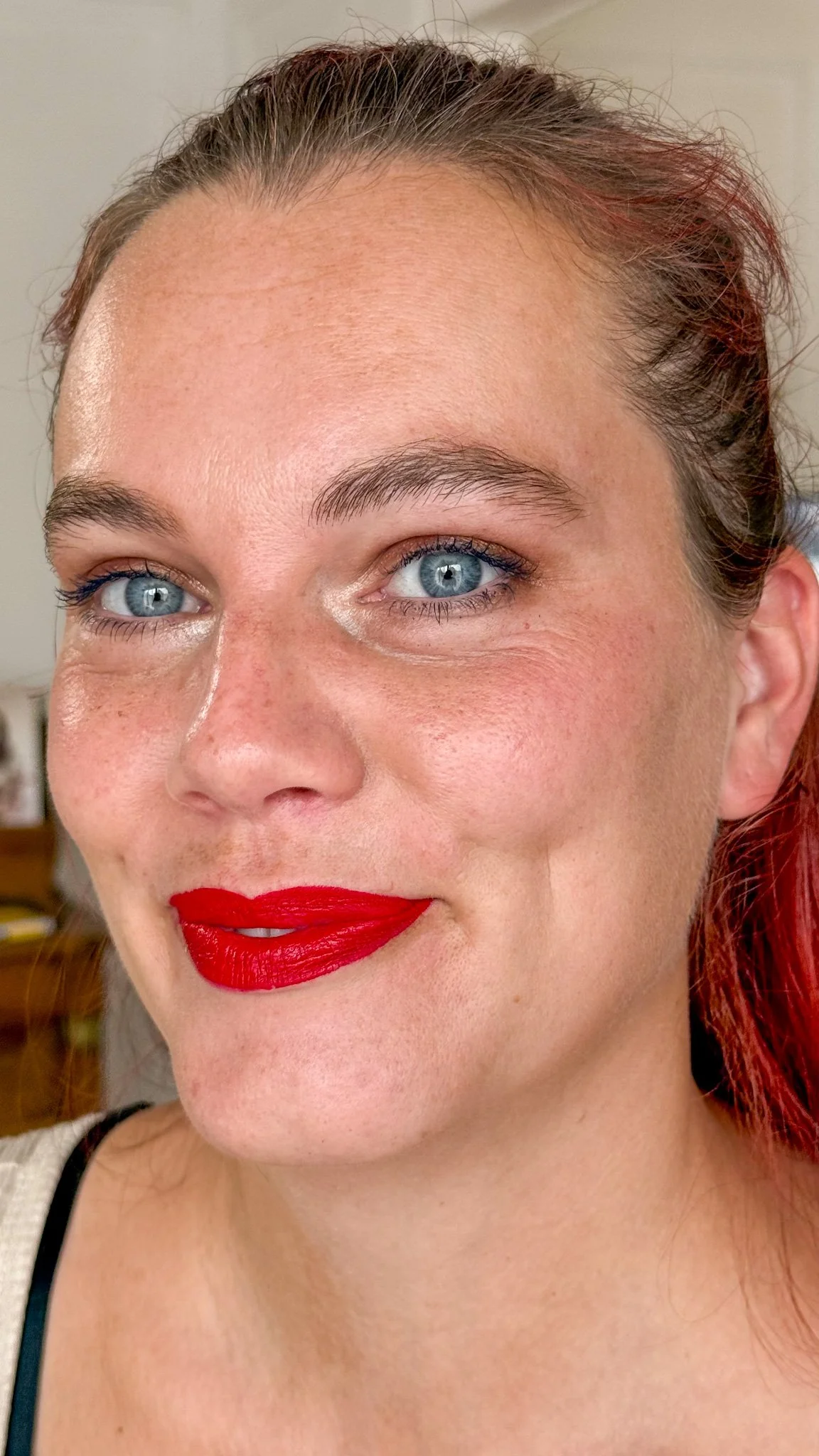 Un gros plan du visage d'une femme avec des cheveux roux, des yeux bleus, portant du rouge à lèvres rouge vif, souriant légèrement.
