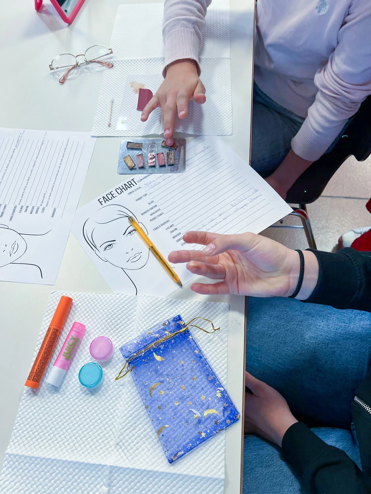 Une table avec des feuilles de papier, des crayons, un plateau avec différentes couleurs de maquillage, un dessin de visage, et un sac en tissu bleu avec des motifs de lune et d'étoiles, ainsi que deux petits pots bleus et un bâton de colle.