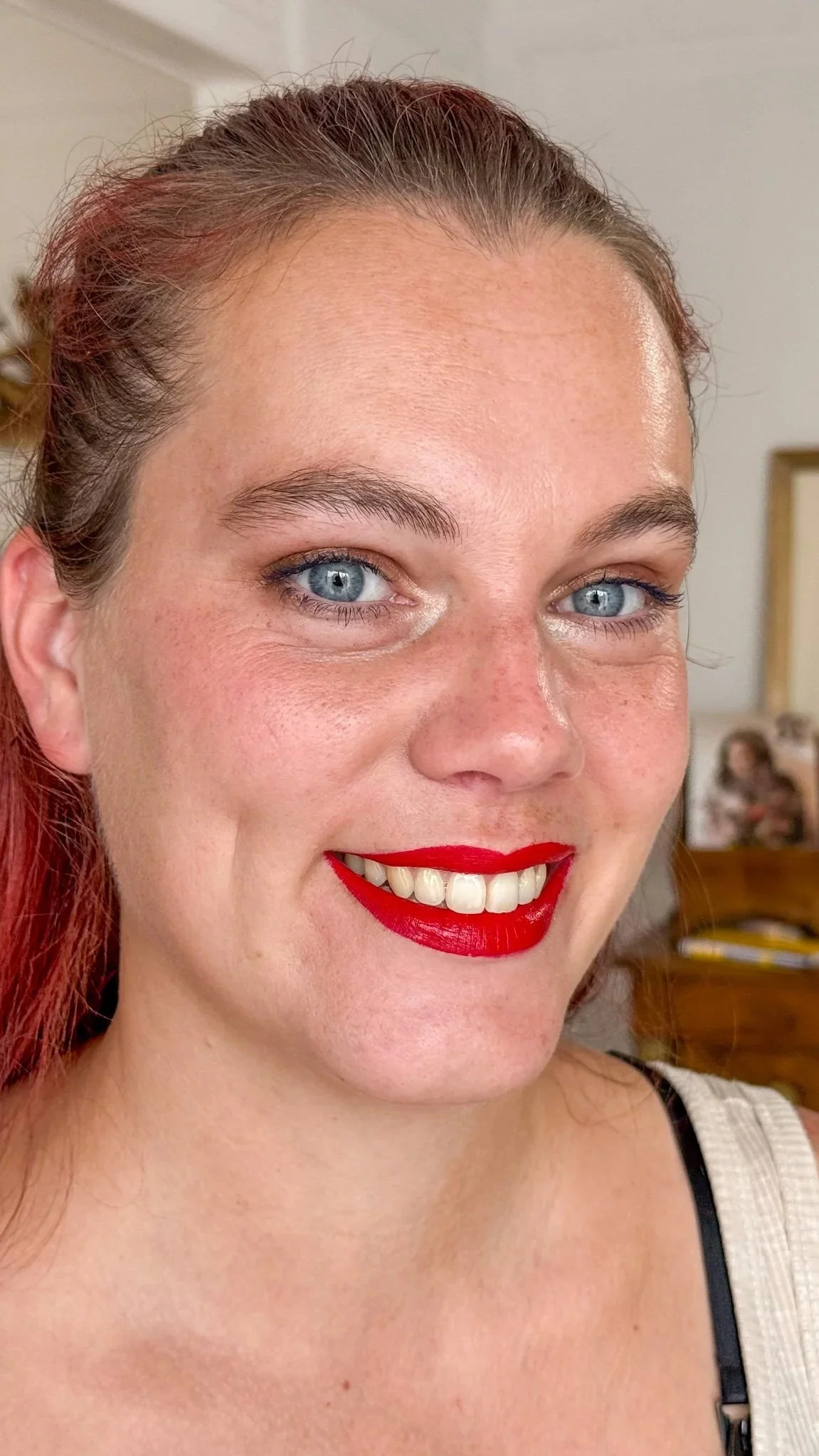 Une femme souriante avec des lèvres rouges, des yeux bleus et des cheveux roux, prise dans un intérieur domestique.