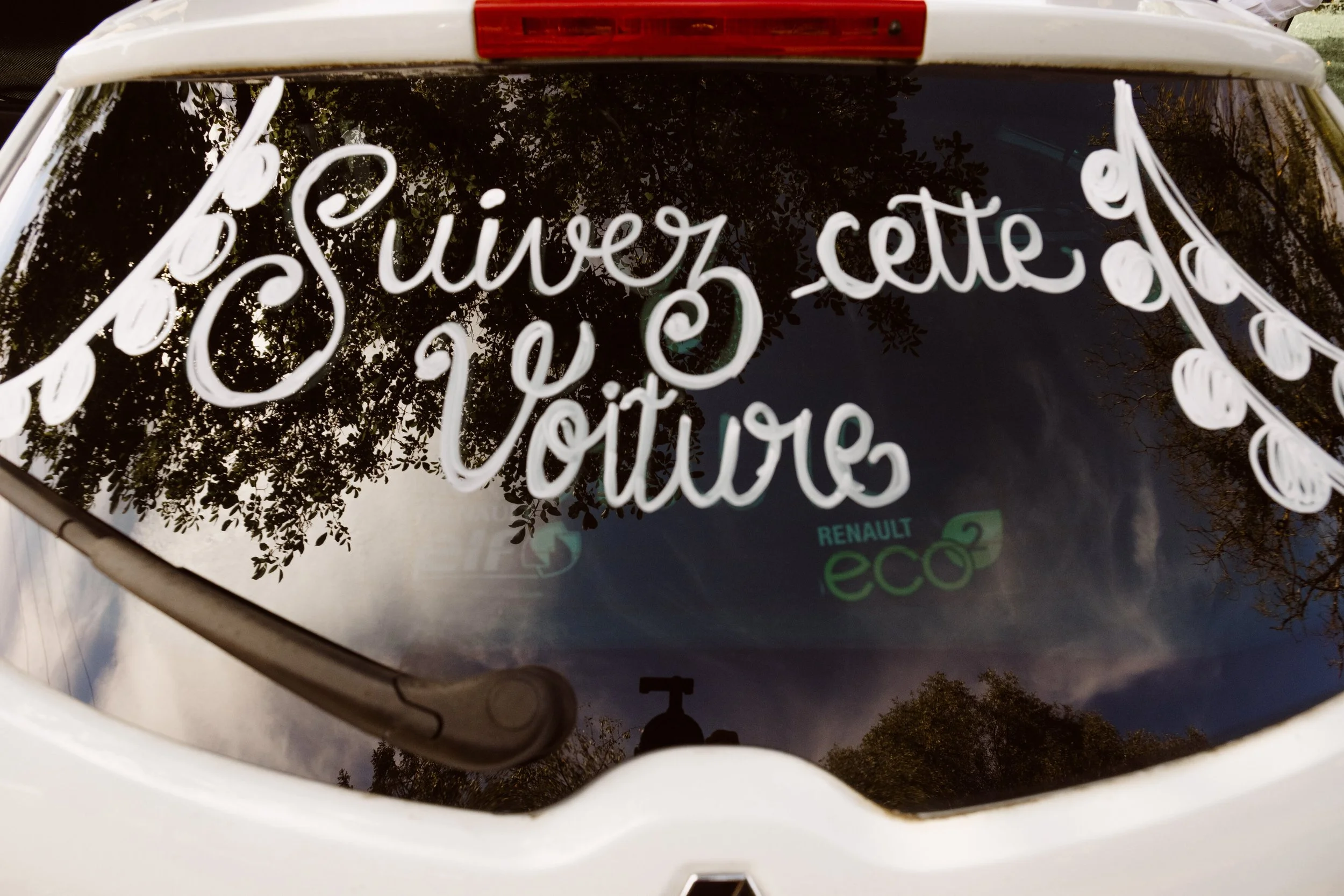 Fenêtre arrière d'une voiture blanche avec une inscription en lettres blanches : "Suisvez cette Voiture", reflet de l'arbre, du ciel et du drapeau "Renault Eco2".