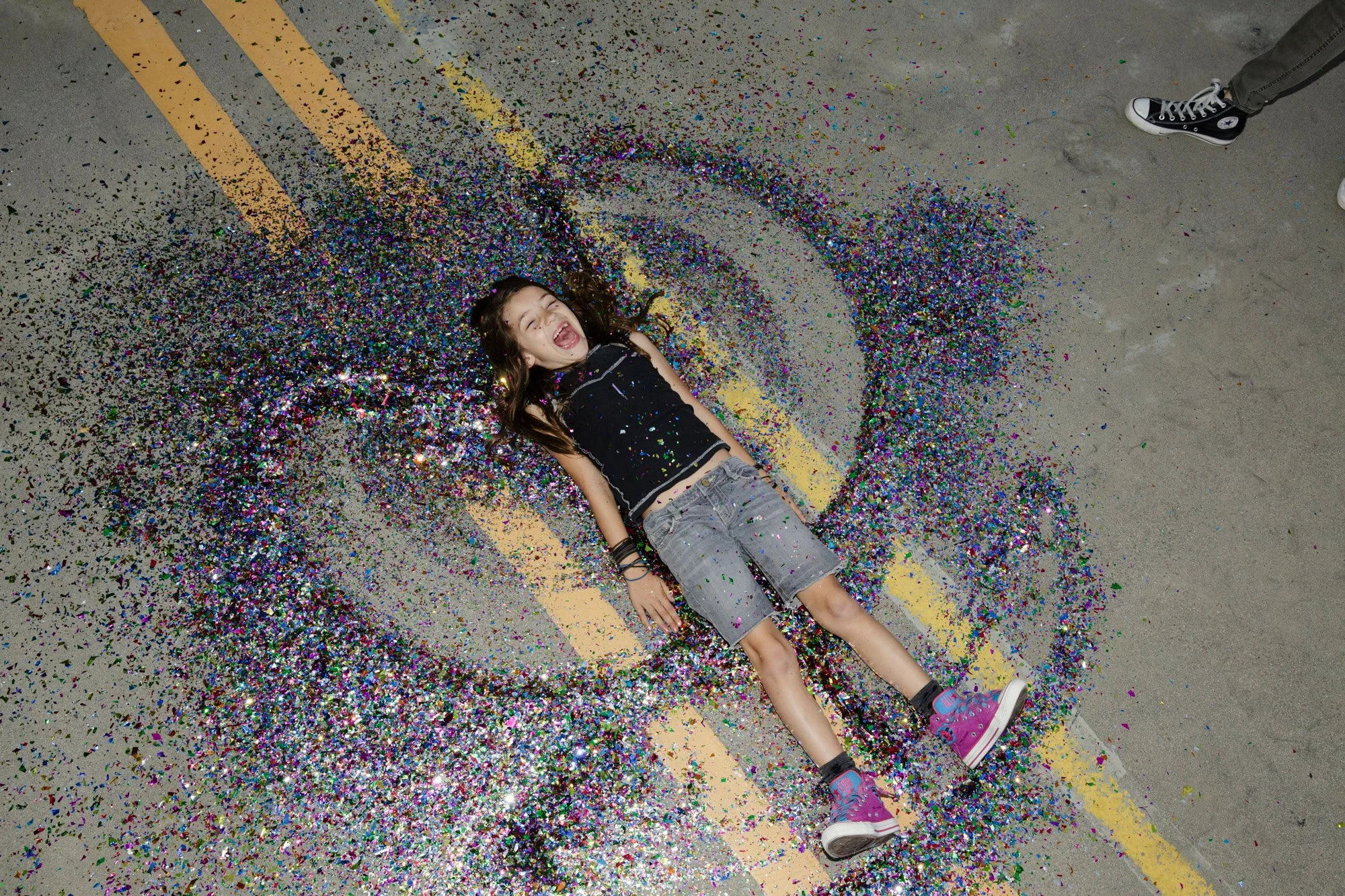 Young girl lying on the ground surrounded by colorful confetti, smiling with arms and legs extended, wearing a black tank top, gray shorts, and purple sneakers, on a street with yellow lines.
