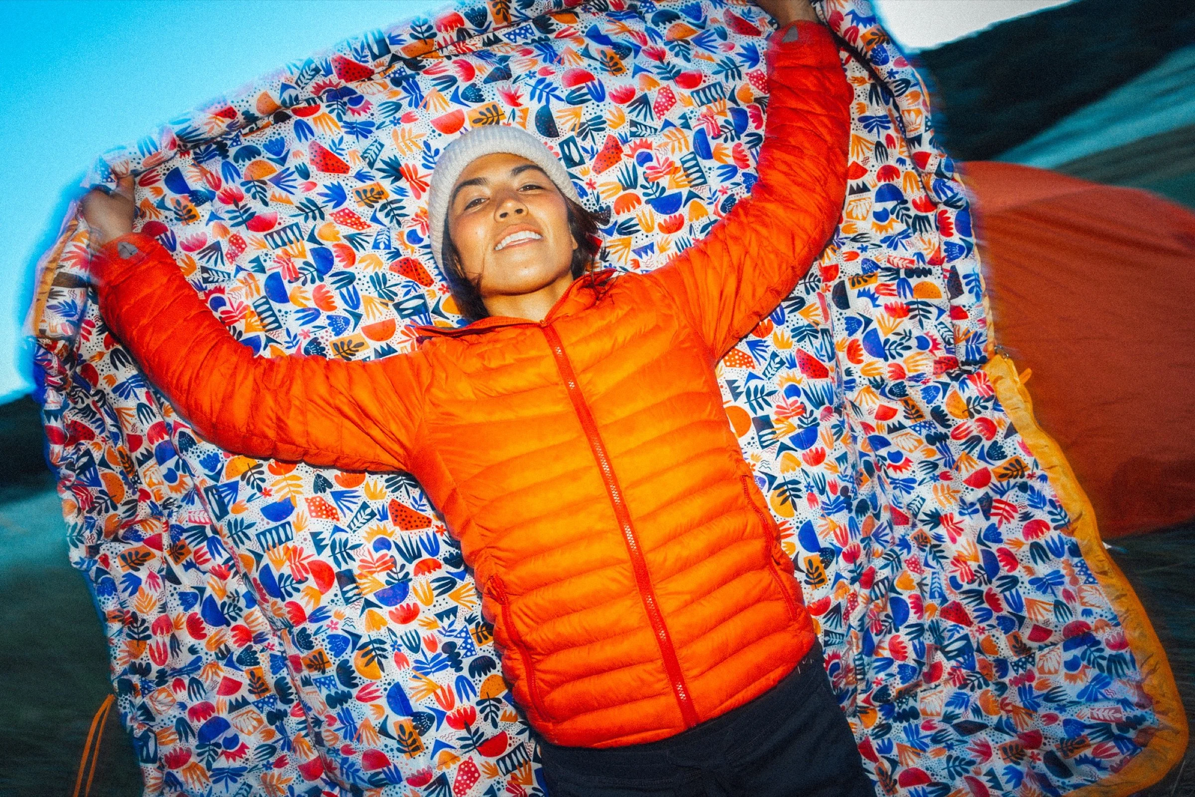 Woman in an orange puffer jacket smiling on a patterned blanket