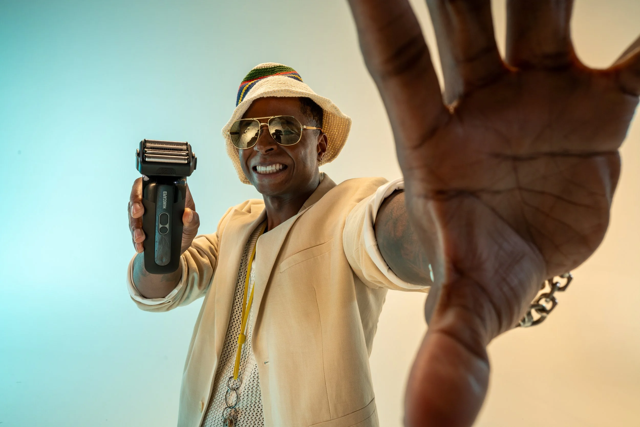 A man wearing sunglasses, a beige blazer, a patterned shirt, a colorful hat, and a white vest is smiling and reaching towards the camera with his hand, which is close to the lens.