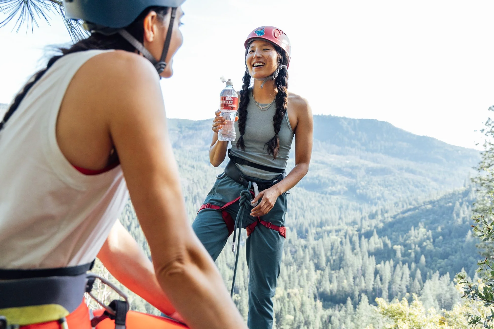 Two women mountain climbing outdoors in a forested mountain landscape, wearing helmets and climbing gear, with one smiling and holding a water bottle.