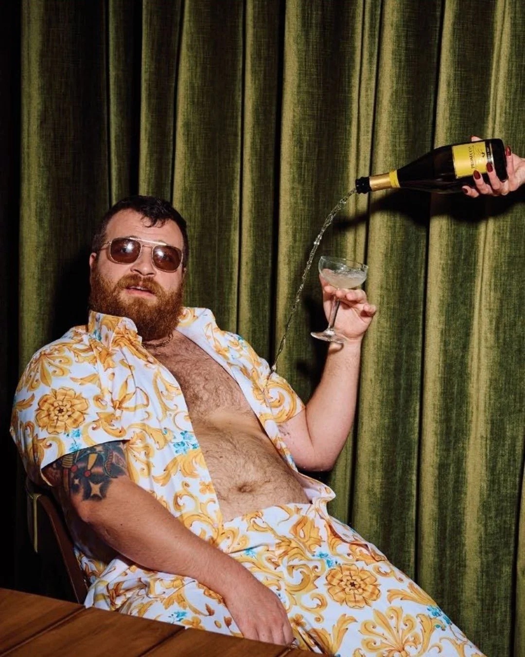 Bearded man in a gold floral shirt holding a martini while champagne pours