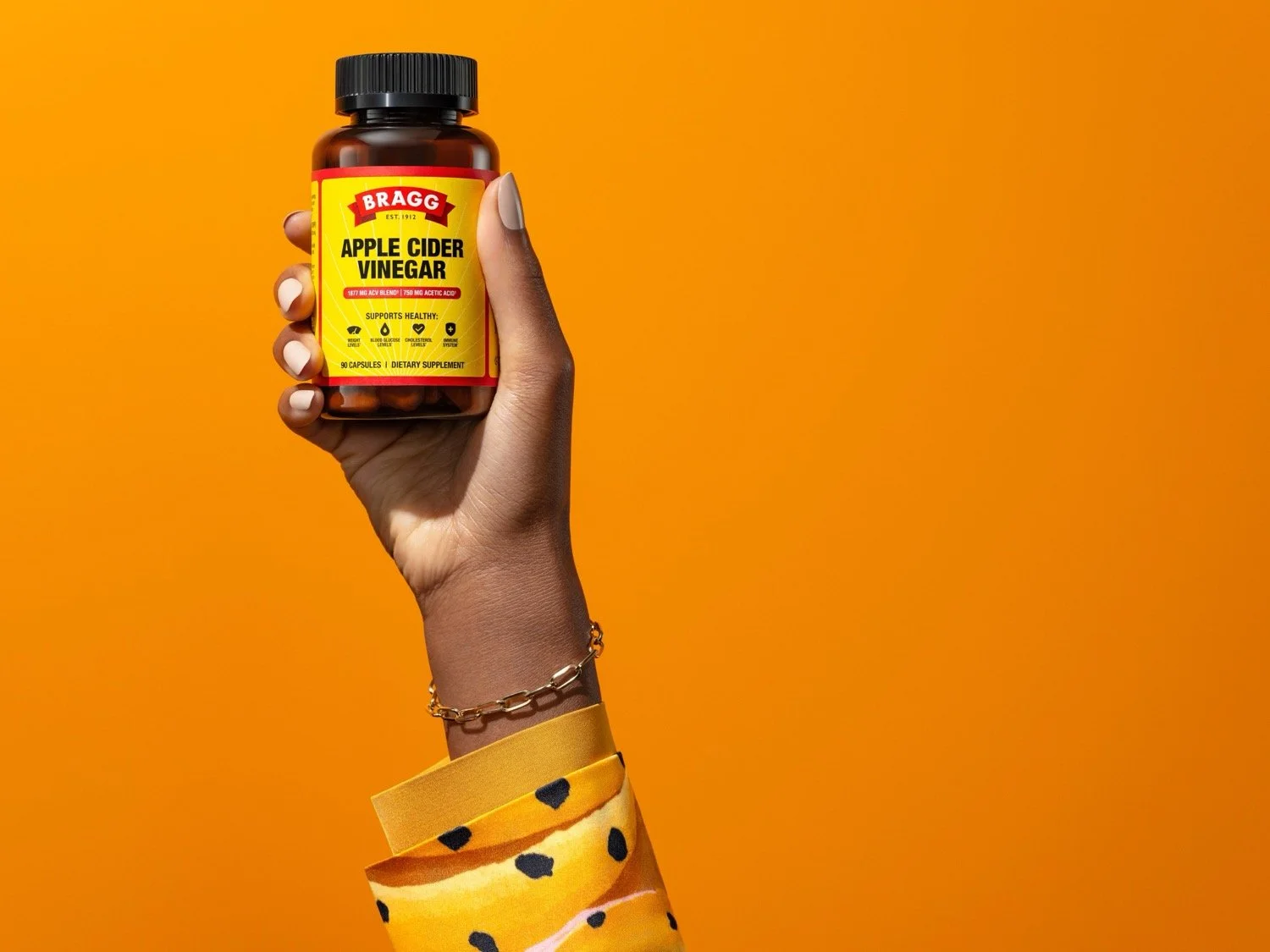 Hand holding up a bottle of Bragg Apple Cider Vinegar against an orange background