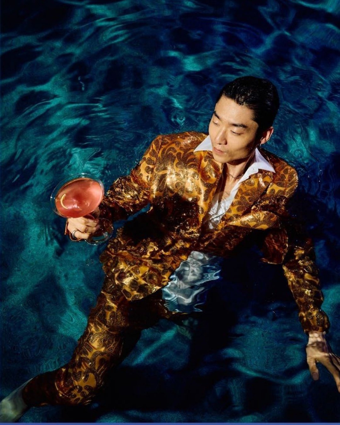 Man in a gold sequined suit floating in a pool holding a cocktail