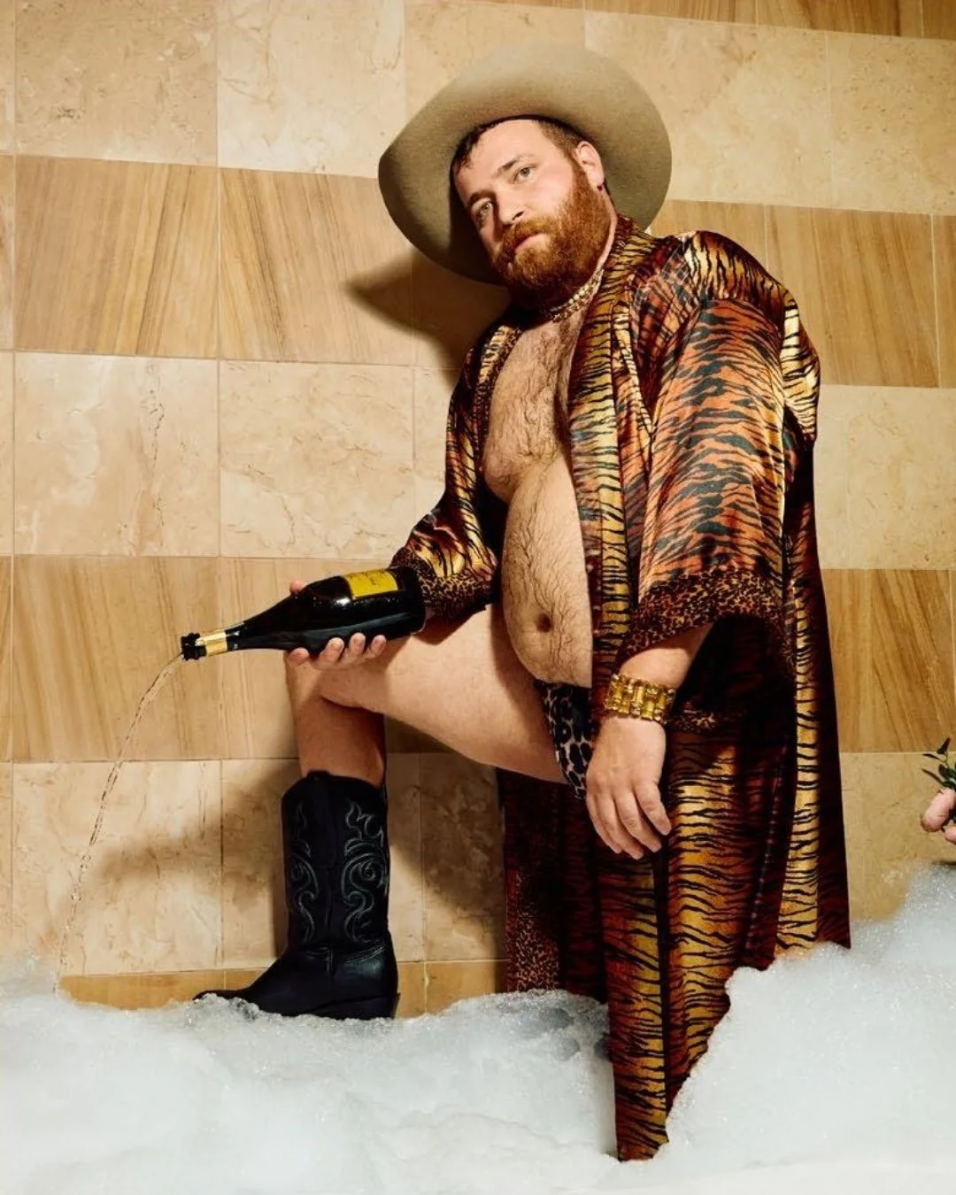 Man in a cowboy hat and tiger-print robe pouring champagne in a bubble bath
