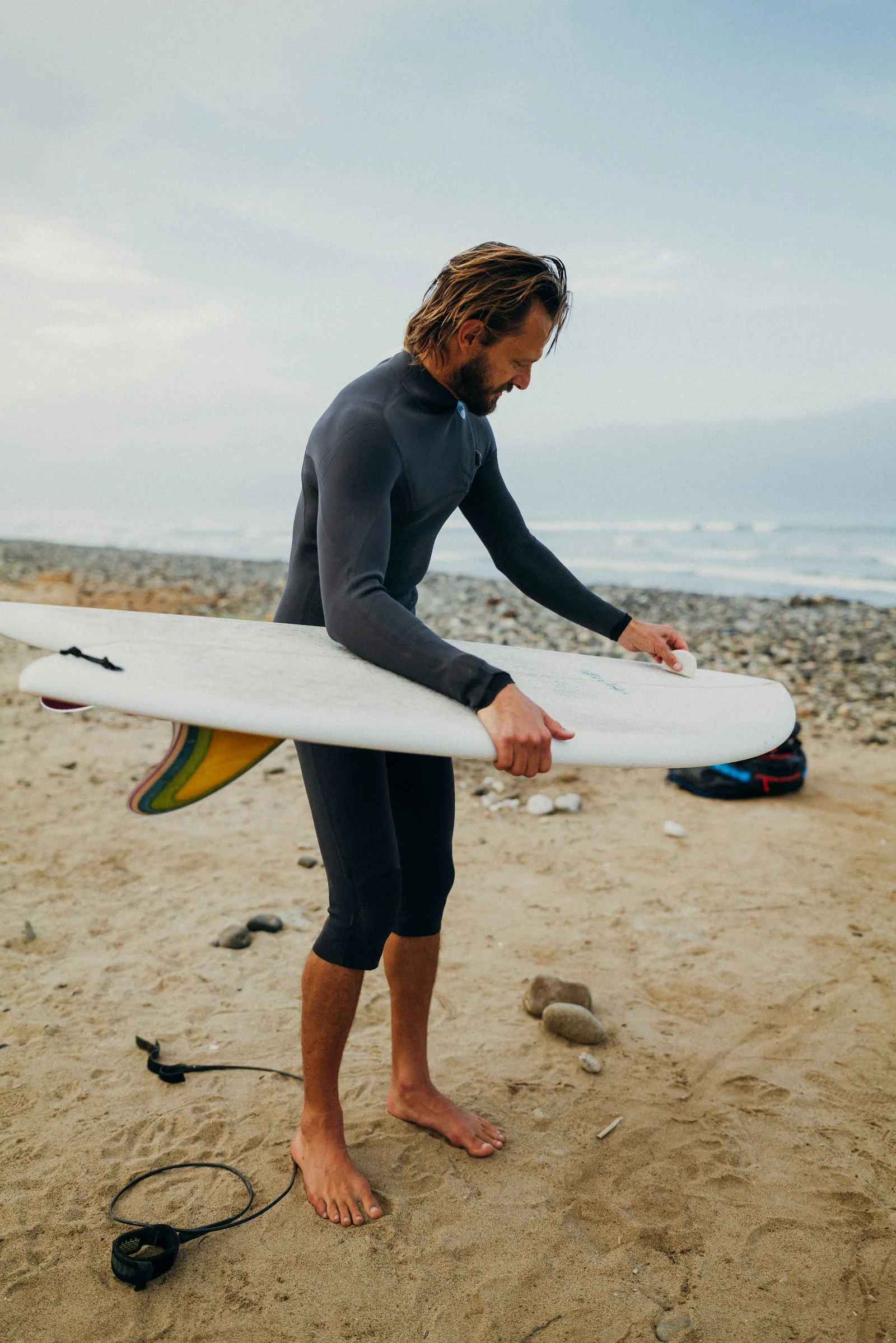 Nick in a wetsuit waxing a surfboard on the beach