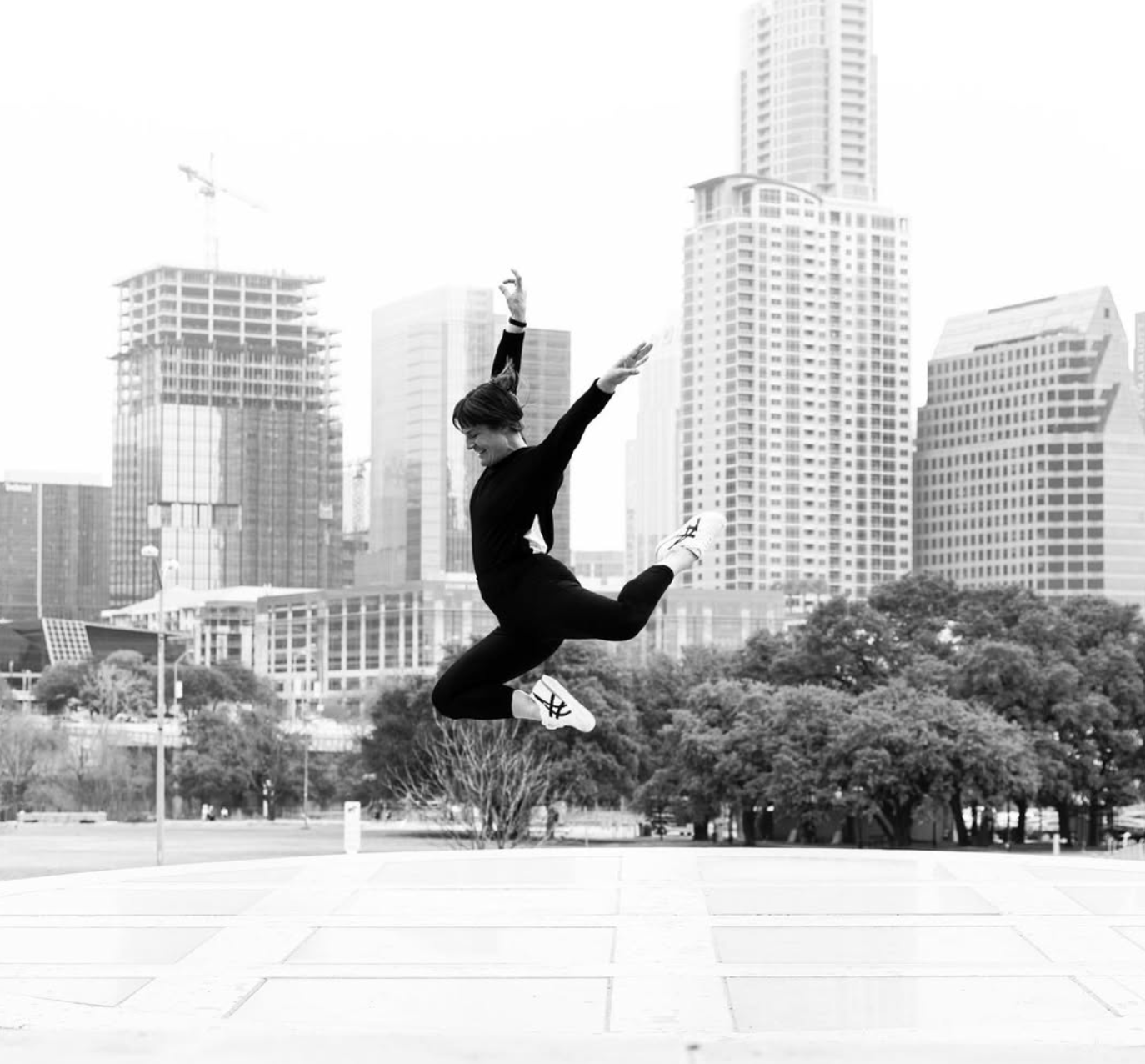 A woman jumping in the air with one leg bent behind her and arms raised in a city park, with skyscrapers and trees in the background.