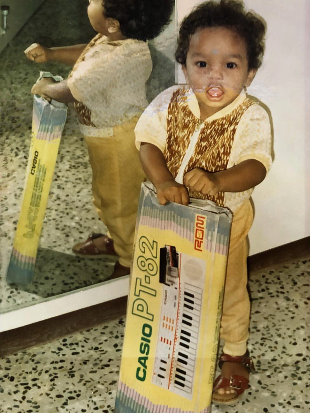 A young boy with curly hair and a patterned shirt holding a boxed Casio PT-82 keyboard, standing on a speckled tile floor indoors.