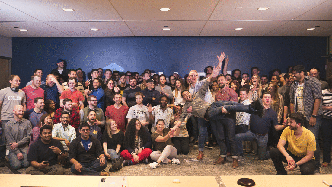 A large group of people gathered indoors, with some seated on the floor and others standing. A man is being lifted horizontally by two people, smiling and waving at the camera. Everyone appears happy and engaged, in a casual setting.