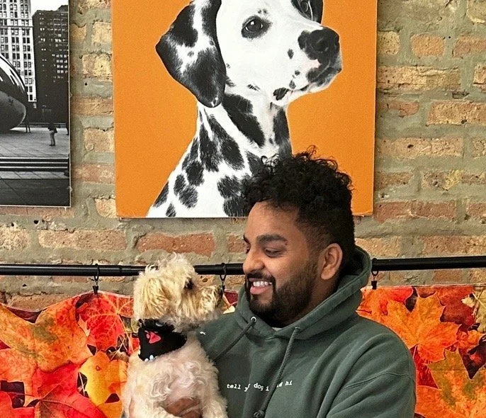 A man with a beard smiling while holding a small dog, sitting on a floral-patterned couch with autumn leaves design. In the background, a wall with a large painting of a Dalmatian dog in black and white on an orange background.