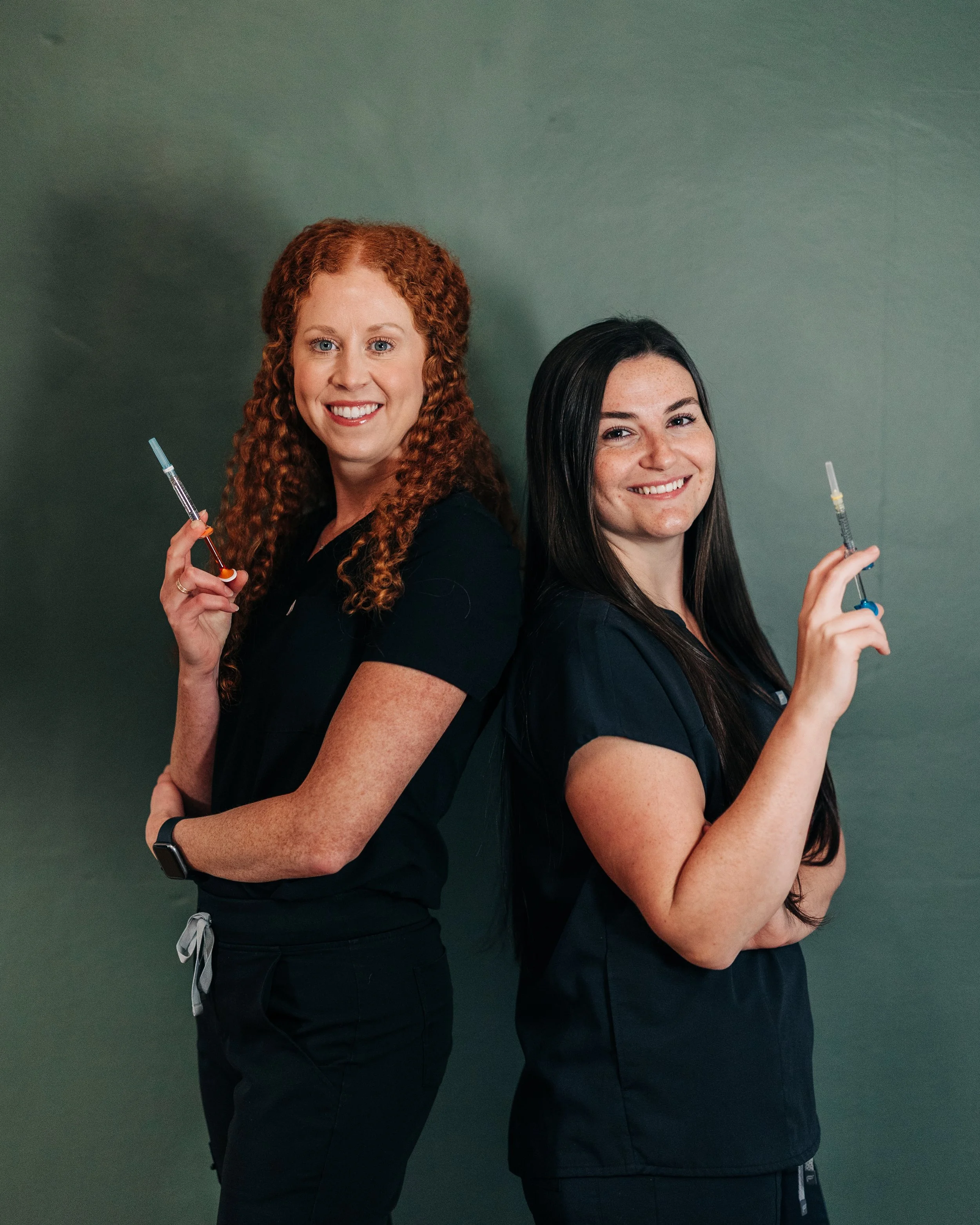 Two smiling women wearing dark scrubs, standing back to back, each holding a syringe, against a plain dark green background.