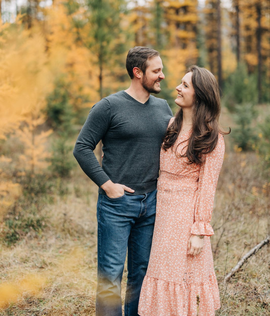 Kaite and Ryan standing close together outdoors among autumn trees, smiling and looking at each other.