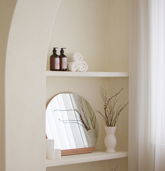 Decorative shelf with two bottles of liquid soap or lotion, three rolled white towels, a round mirror, and two vases with branches, all set against a light-colored wall with white curtains.