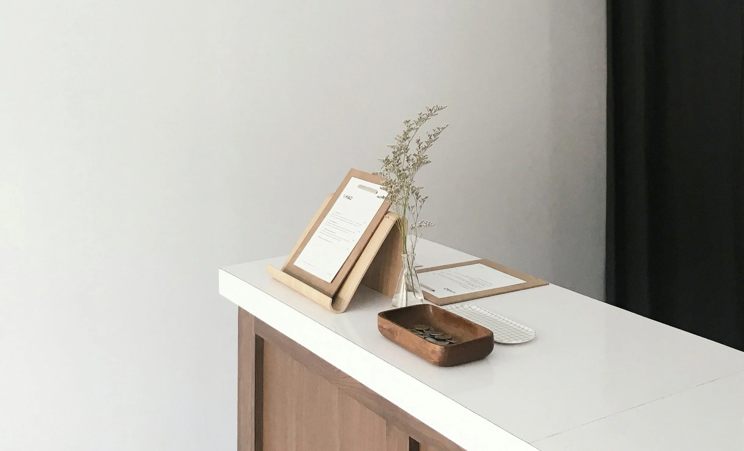 Minimalist reception desk with a small vase of dried flowers, a framed menu, a wooden tray with coins, and a clipboard with papers.