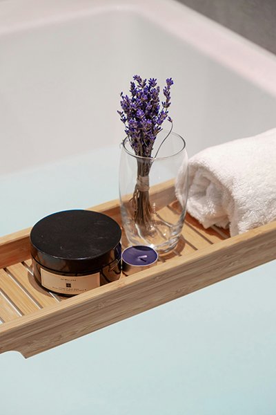 A wooden tray containing a glass vase with lavender, a white rolled towel, a black candle, and a small purple candle.