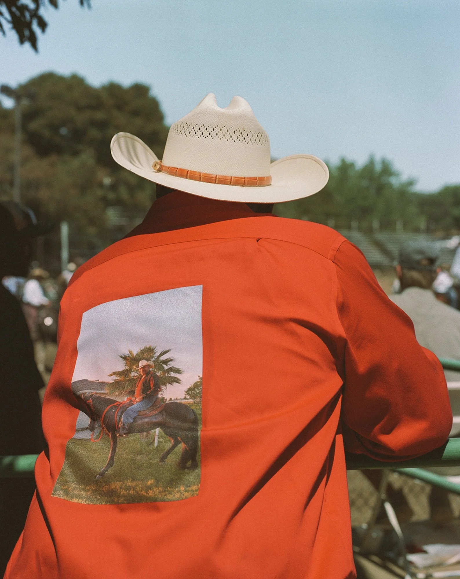 Person wearing a white cowboy hat and an orange jacket with a horse riding picture on the back, outdoors.