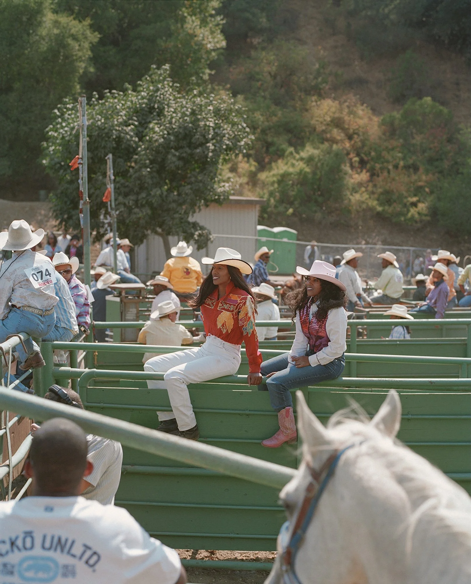 Two women sitting on a green cow pen at a rodeo, wearing cowboy hats and western attire, with a crowd of people and trees in the background.
