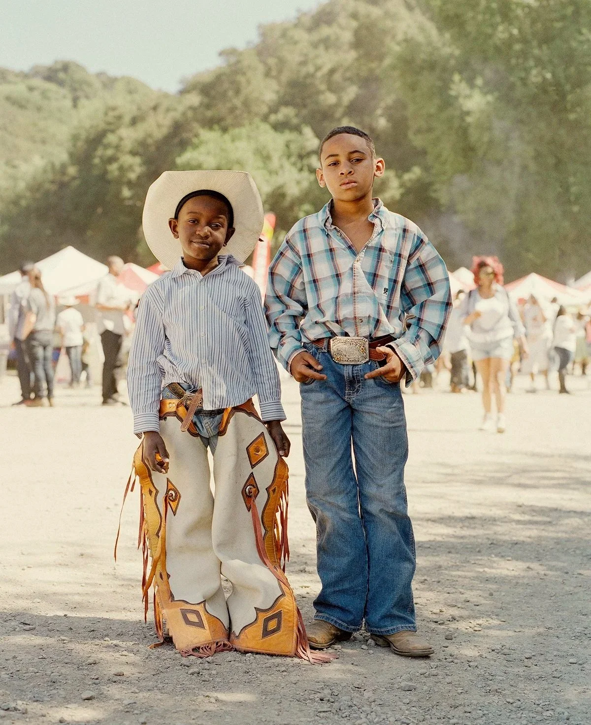 Two young boys standing together at an outdoor event or festival, with tents and people in the background. One boy is wearing a cowboy hat, a striped shirt, and large decorated pants, while the other is dressed in a plaid shirt and jeans with a large