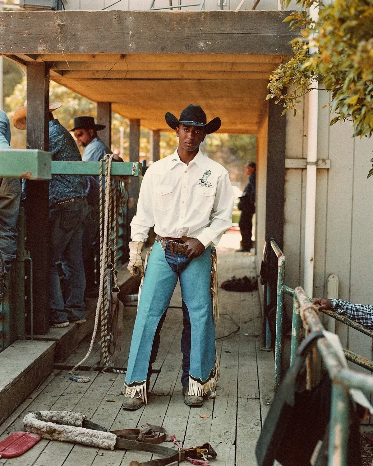 A young man dressed as a cowboy in a white shirt and blue chaps, standing on a wooden deck with other people in the background, some wearing cowboy hats.
