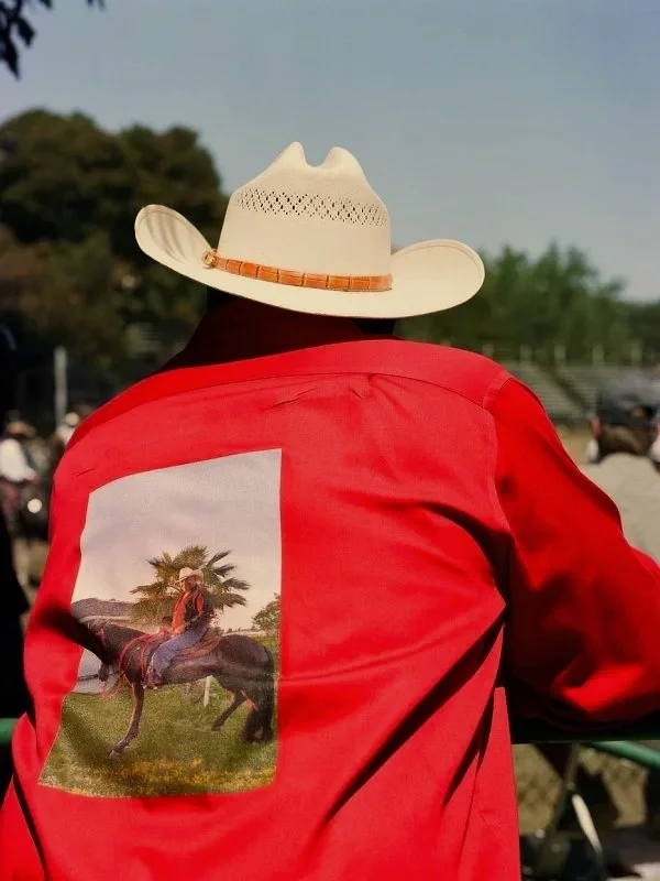 A person wearing a wide-brimmed cowboy hat and a red jacket with a large patch of a man riding a horse in a grassy field.