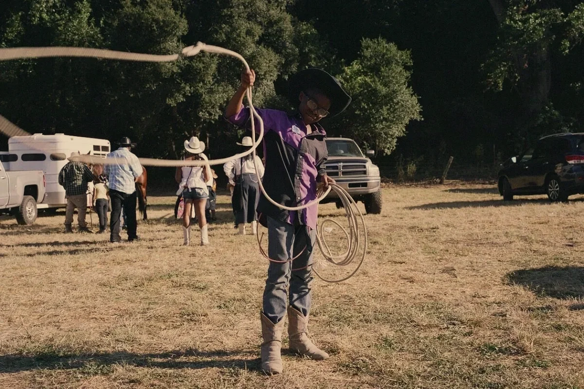 A person dressed as a cowboy, wearing a cowboy hat, glasses, and a purple and black shirt, is holding a lasso or rope, standing in a field with other people and vehicles in the background.