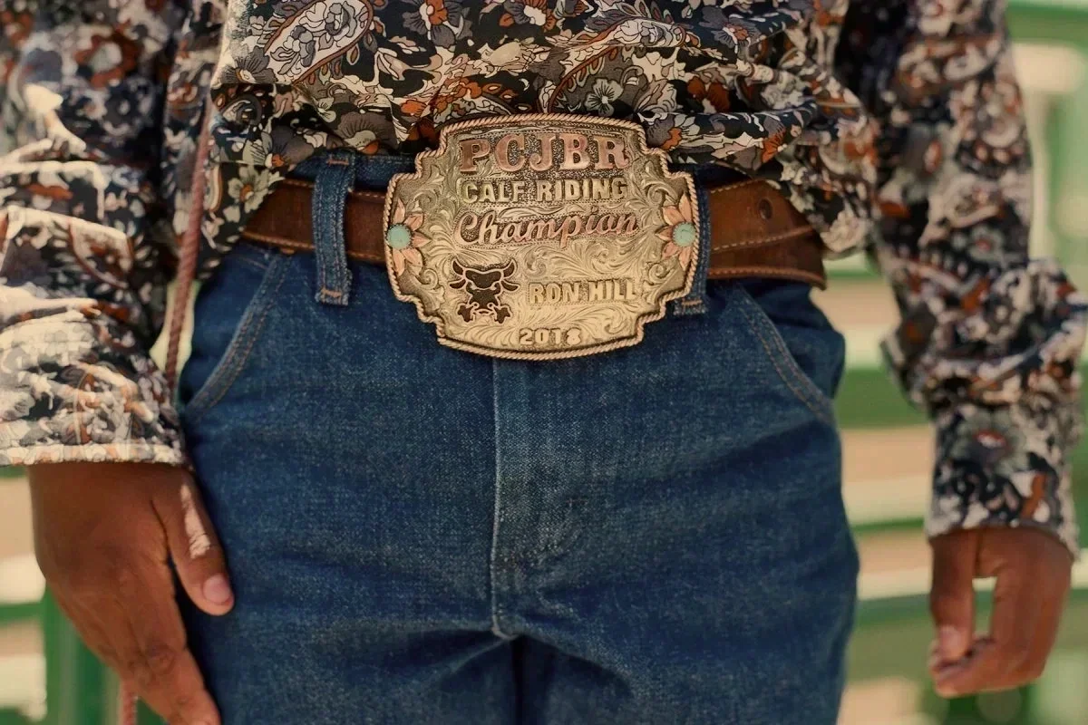 Close-up of a person's waist wearing blue jeans and a large decorative belt buckle that reads 'POJBR Calf Riding Champion 2018' with an image of a bull's head.
